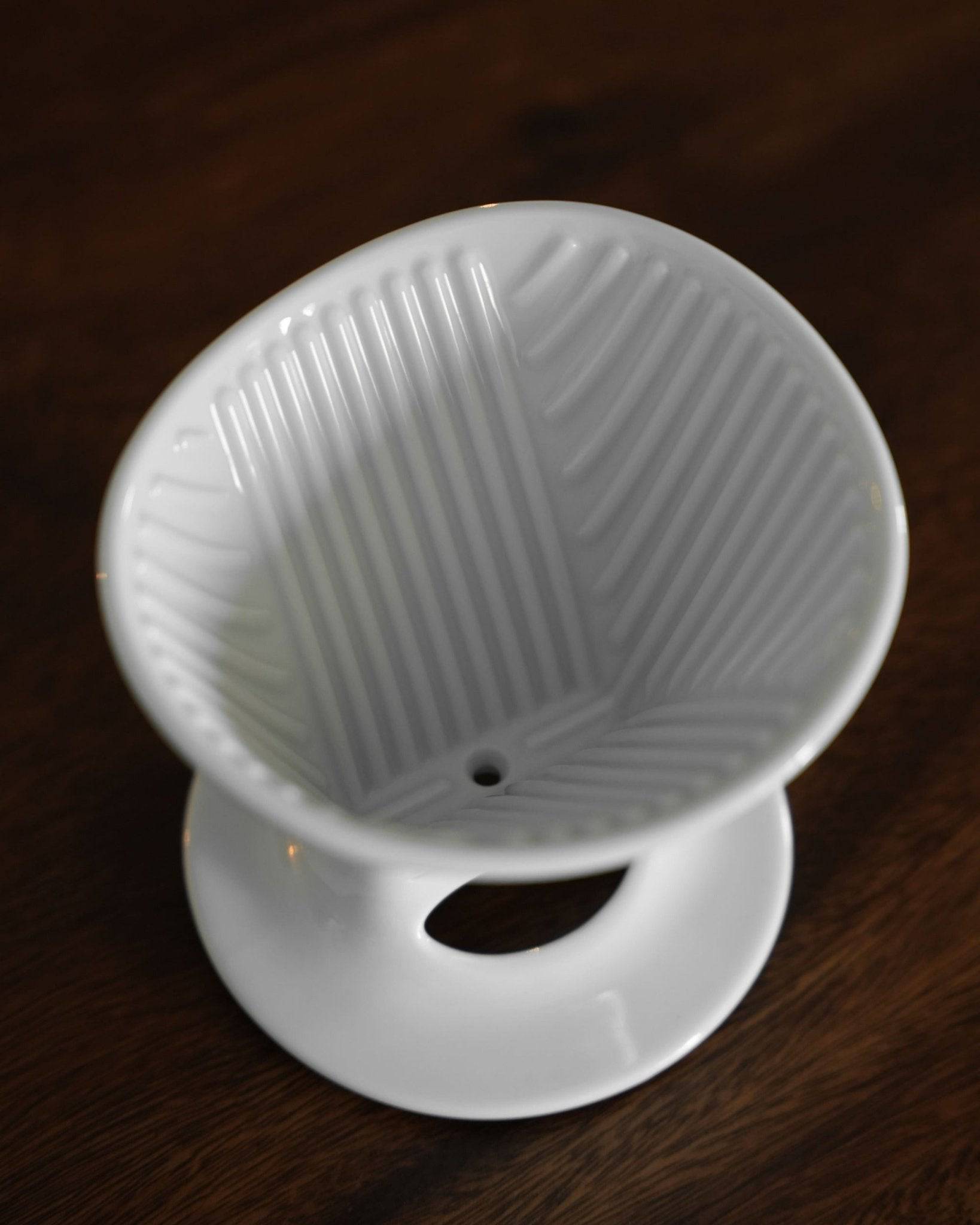 White ceramic pour-over coffee dripper with distinctive ribbed interior design, single hole at bottom, and attached saucer, showcasing Melitta and Hasami collaboration on dark wooden surface