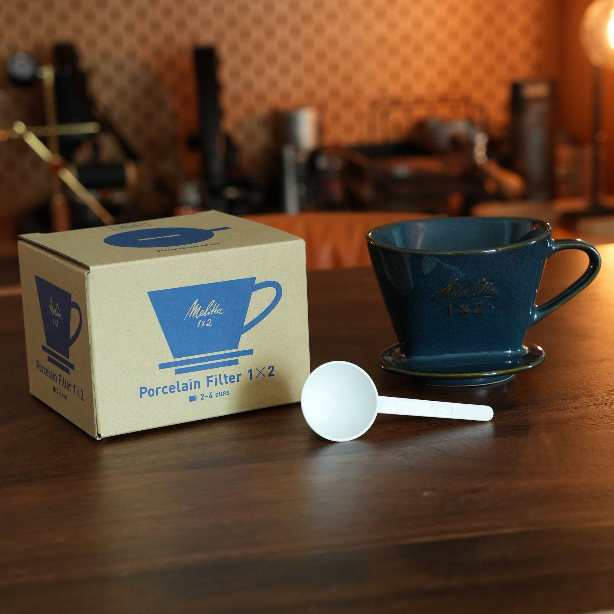 Melitta turquoise blue porcelain pour-over coffee dripper with box and measuring spoon on wooden table, showcasing Japanese craftsmanship and unique fan-shaped single-hole design for optimal brewing