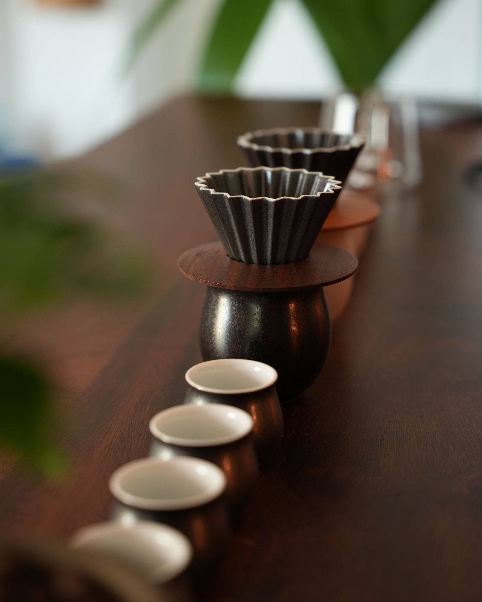Elegant Ninety Plus coffee brewing set featuring ribbed black drippers, wooden stand, and small white tasting cups on a dark wooden surface, with green leaves in foreground, showcasing premium pour-over coffee experience