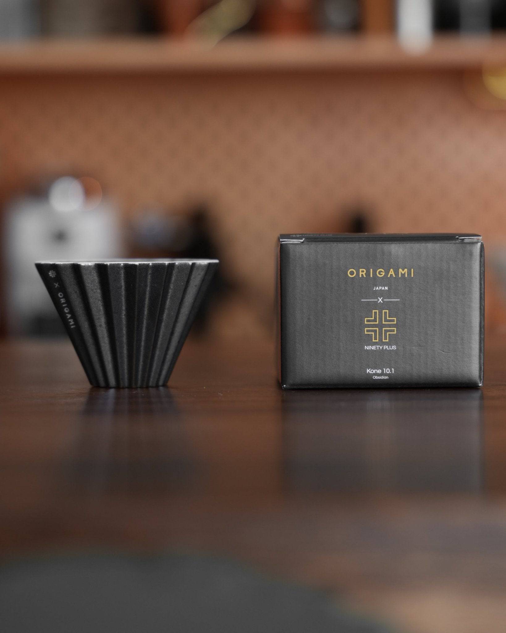 Ninety Plus Coffee Kone 10.1 mini brew dripper and its packaging box, featuring a black folded cone-shaped dripper and a sleek black box with gold Origami and Ninety Plus logos, displayed on a wooden surface in a kitchen setting.