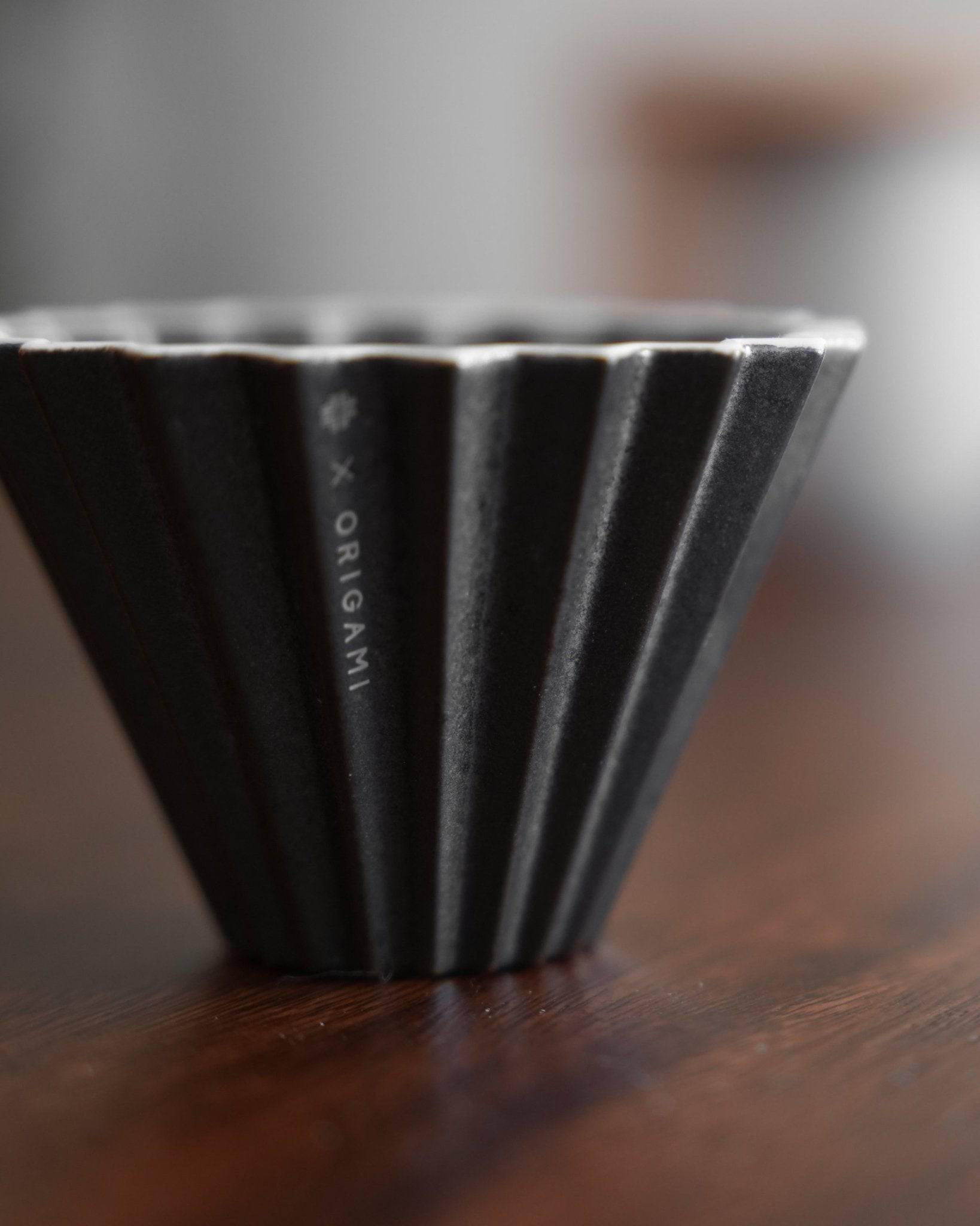 Close-up of Ninety Plus Coffee Kone 10.1 dripper, black obsidian color, ribbed conical design, sitting on wooden surface, with Origami branding visible on side, showcasing elegant minimalist aesthetic for specialty coffee brewing