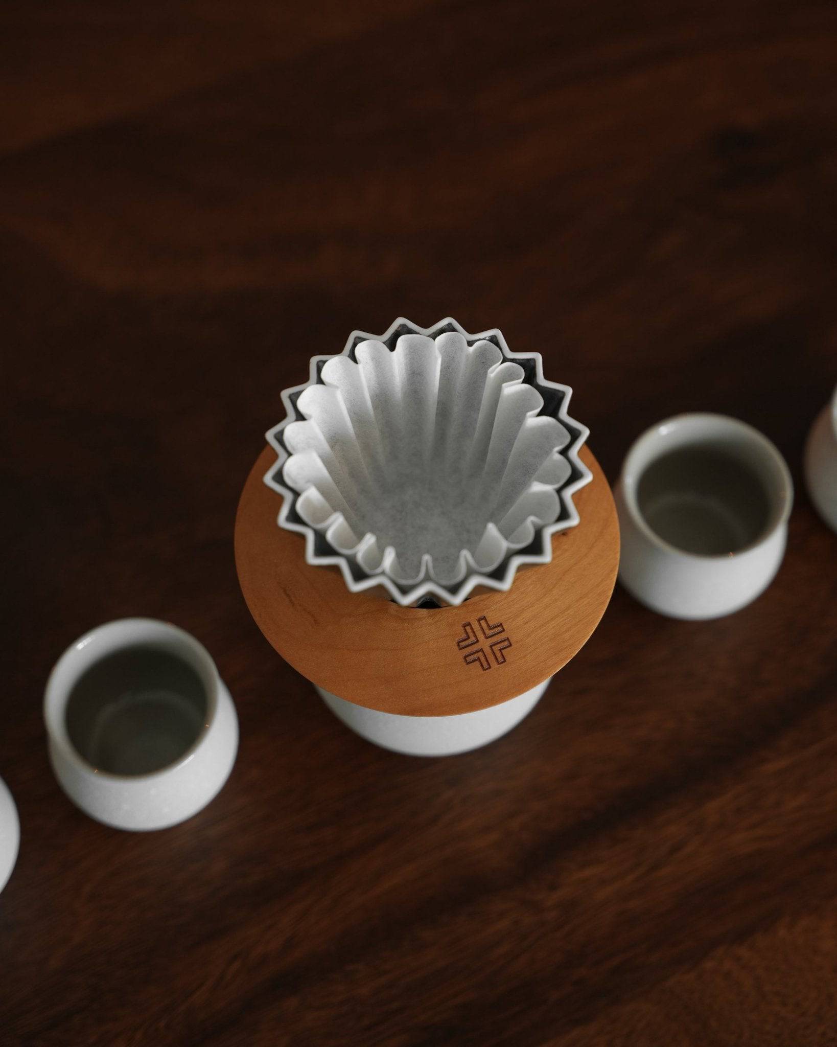 Ninety Plus Coffee Kone Filter 10.1 flat-bottom coffee filter paper set on wooden stand, surrounded by ceramic cups on dark wooden surface, highlighting premium quality and design for precision coffee brewing