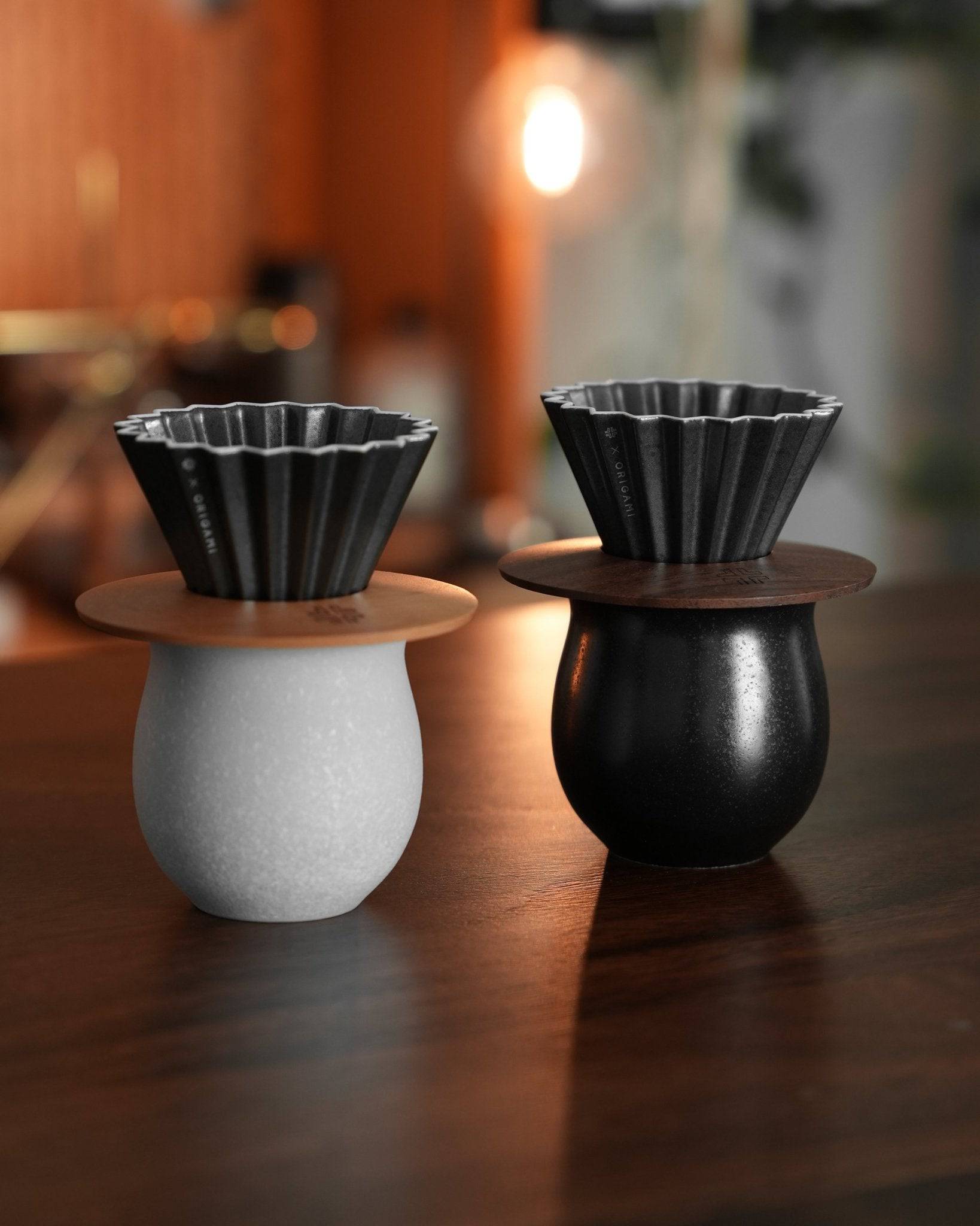Two Ninety Plus Coffee Savour Cups, one white and one black, with fluted pour-over filters on top, placed on a wooden surface in a warm, softly lit environment