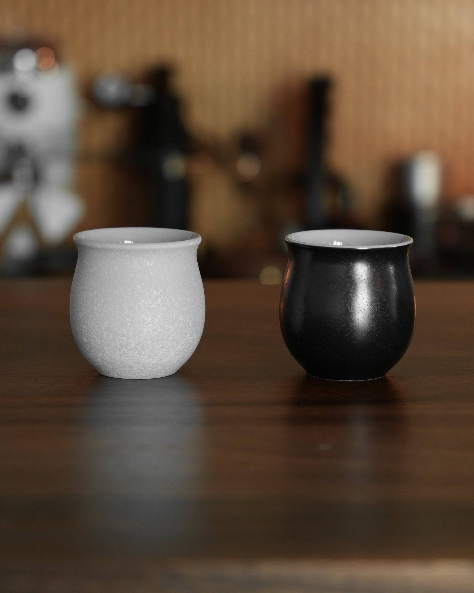 Ninety Plus Coffee Savour Cup 10.1 in white and black, inspired by basalt and obsidian, on wooden surface with blurred coffee equipment background. Elegant, curved design for direct extraction and pure coffee enjoyment.