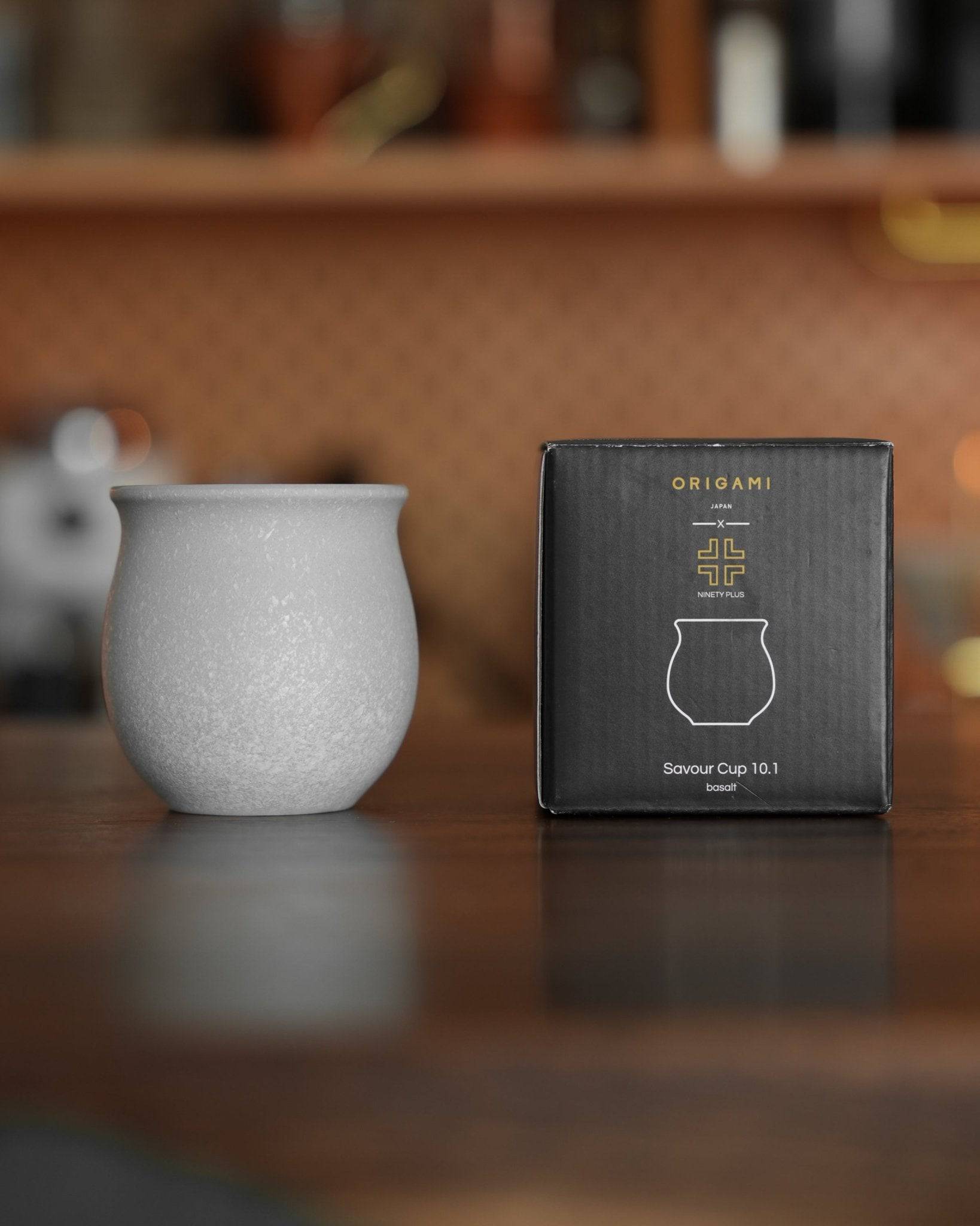 Ninety Plus Coffee Savour Cup 10.1 in basalt white, displayed next to its Origami branded packaging on a wooden surface, showcasing the cup&