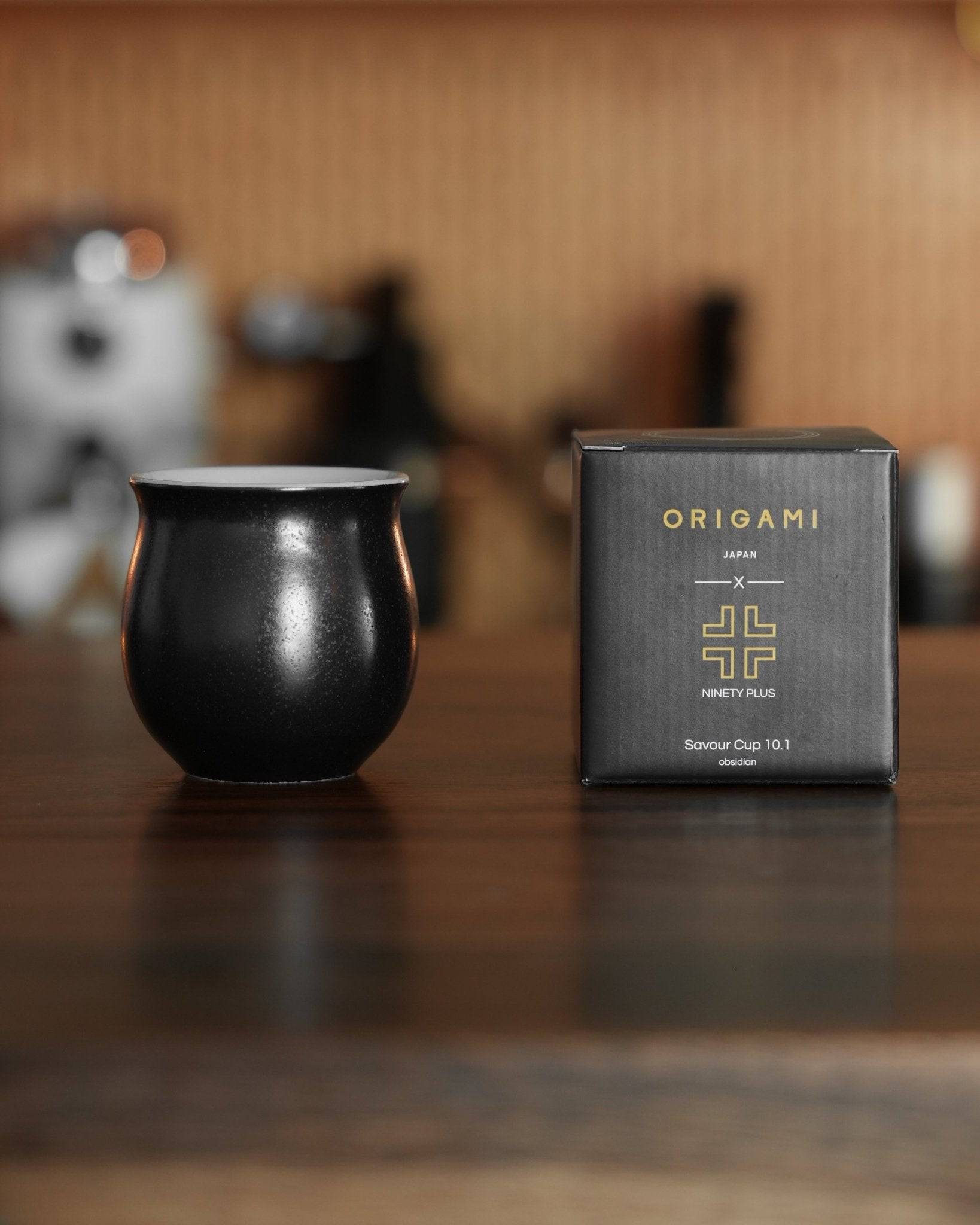 Ninety Plus Coffee Savour Cup 10.1 in obsidian black, displayed next to its Origami Japan packaging box on a wooden surface, with coffee equipment blurred in the background