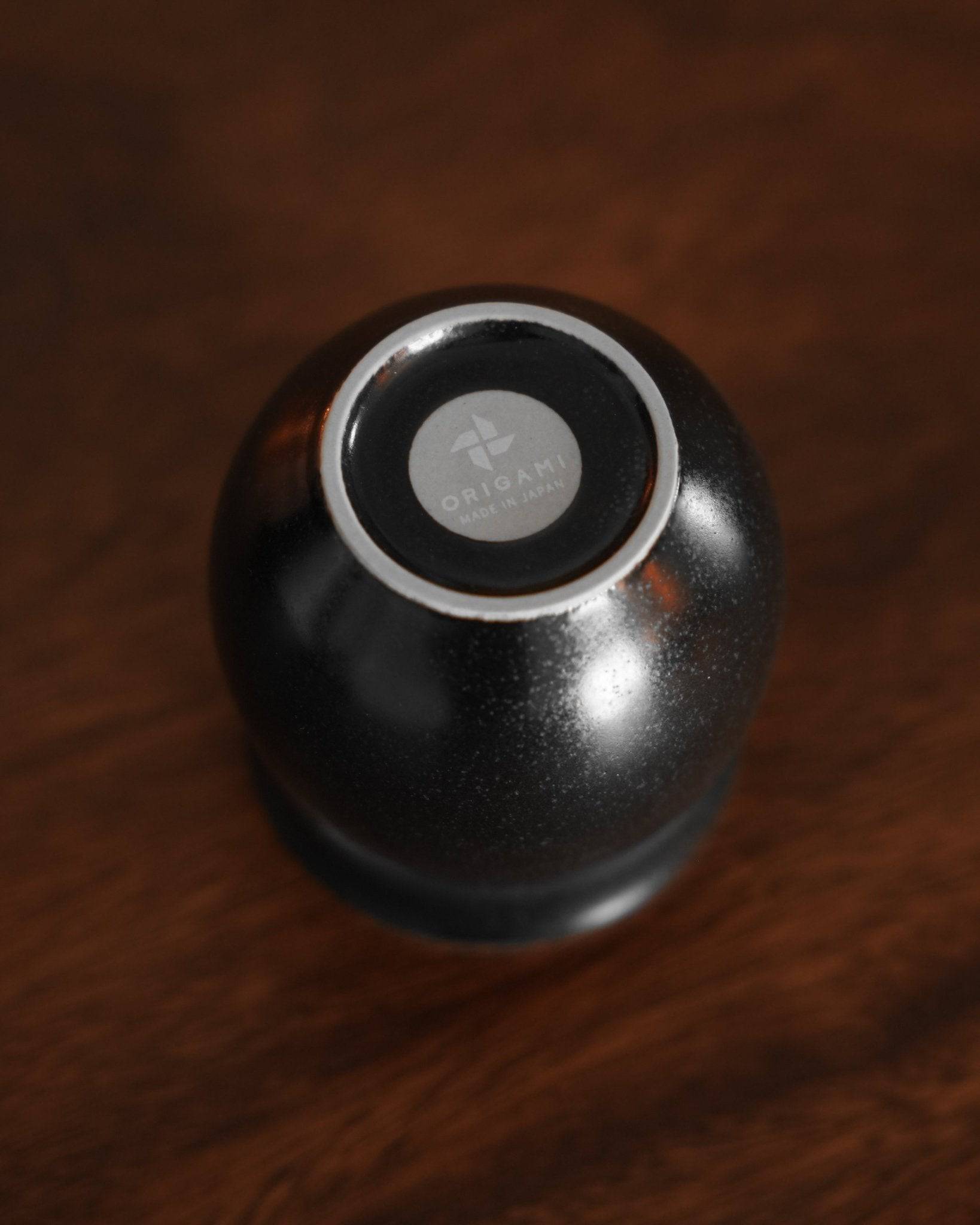 Ninety Plus Savour Cup in obsidian black, showcasing its sleek design and Origami logo on the bottom, placed on a dark wooden surface, highlighting its premium quality and aesthetic appeal for coffee enthusiasts