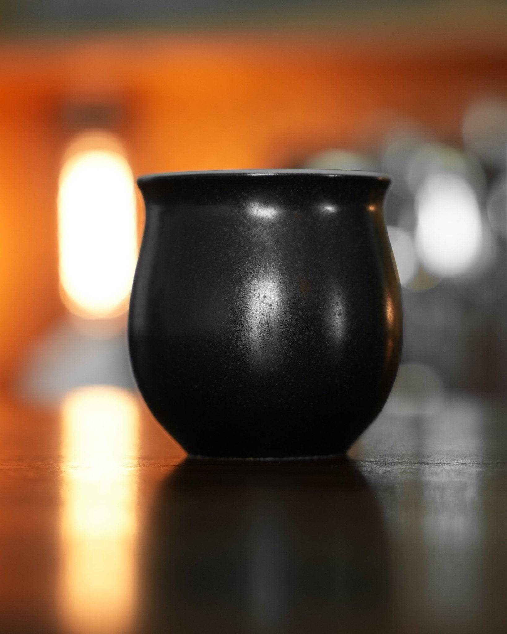 Ninety Plus Coffee Savour Cup in obsidian black, showcasing its sleek design inspired by volcanic rock. Soft orange glow in background highlights the cup&