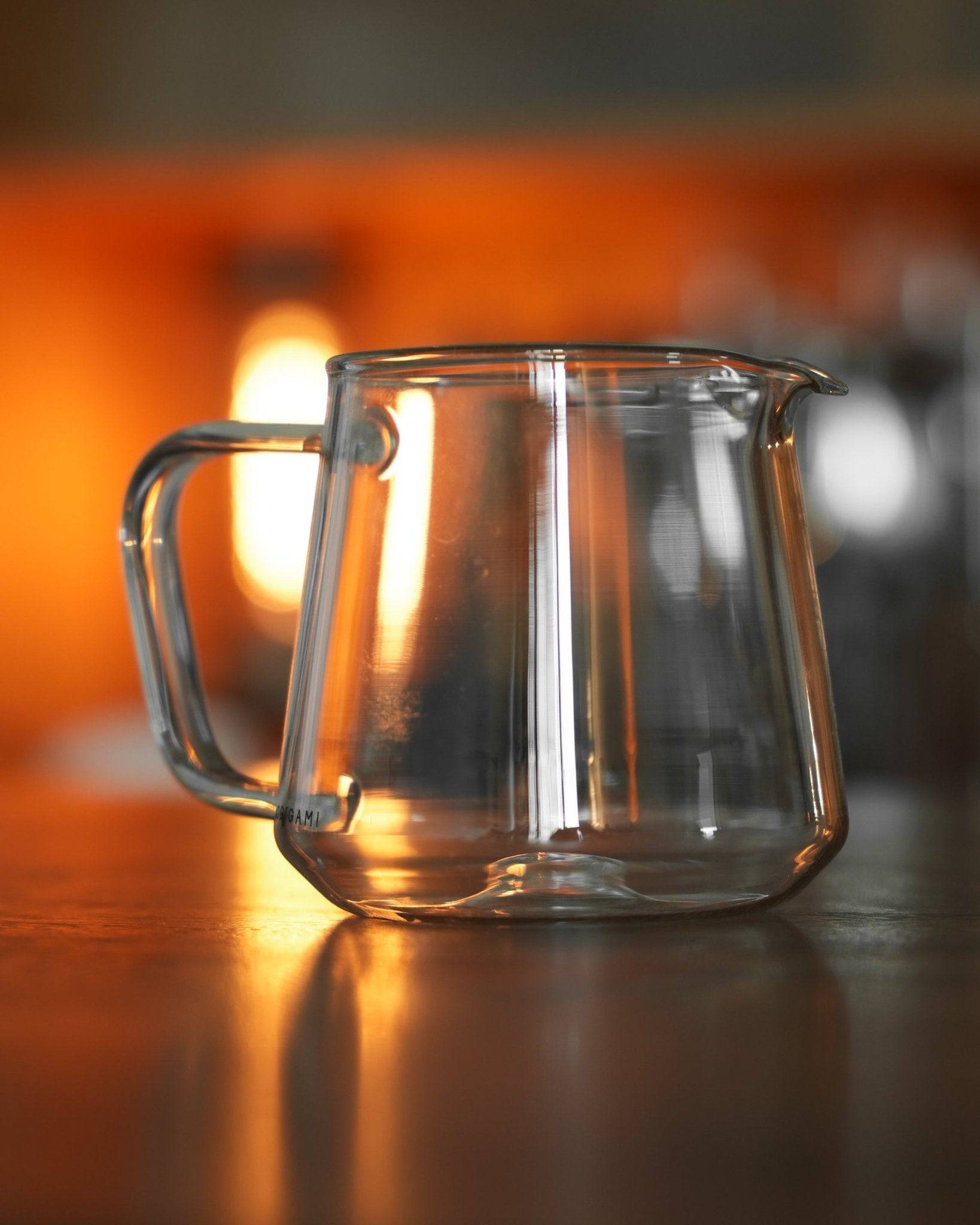 Clear glass coffee serving pitcher with elegant handle, illuminated by warm ambient lighting, reflecting Ninety Plus Coffee&