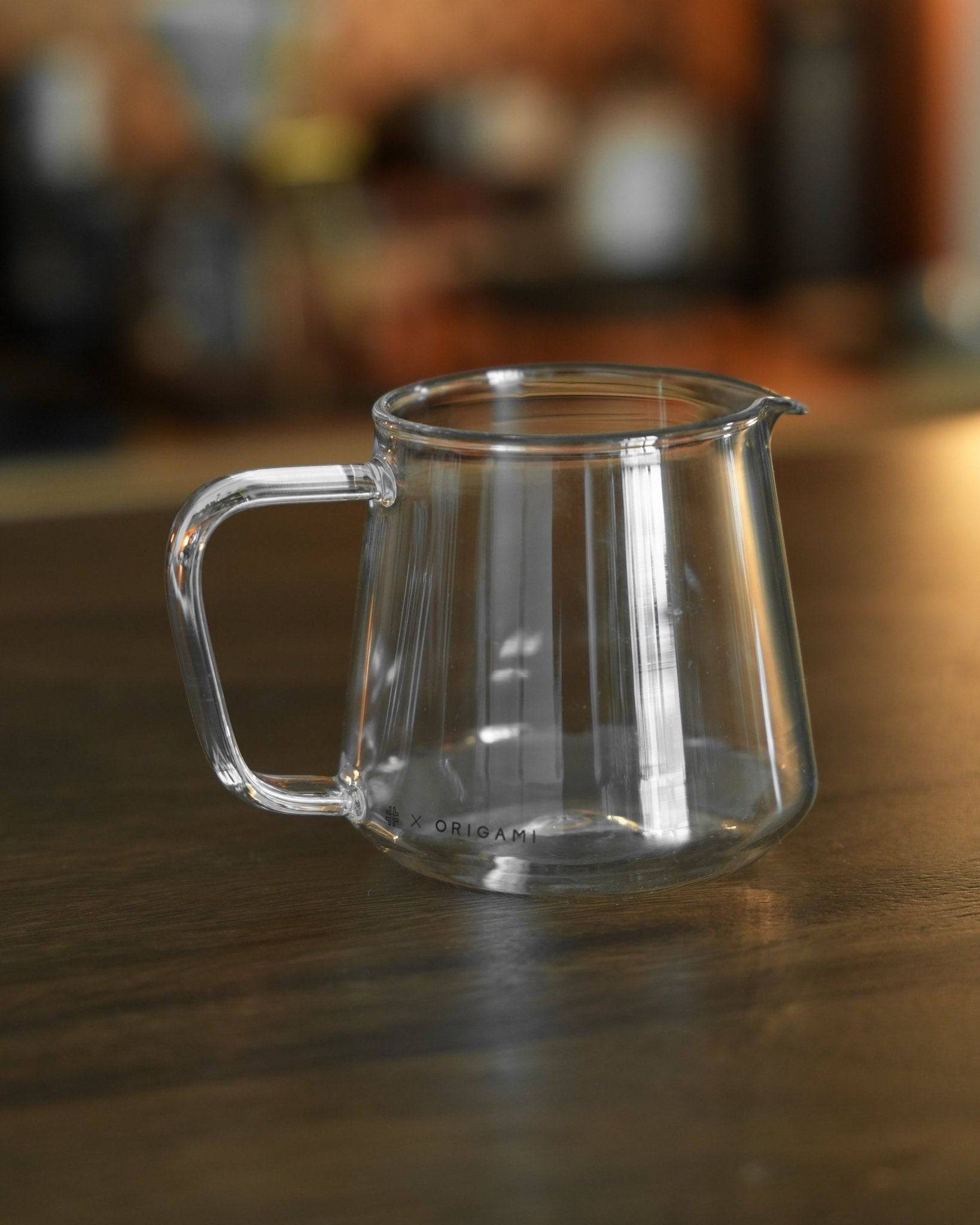 Transparent glass Ninety Plus Coffee Serving Pitcher 10.1 on wooden surface, designed for precise coffee distribution and pairing with Origami filters, showcasing elegant craftsmanship for premium coffee sharing experiences