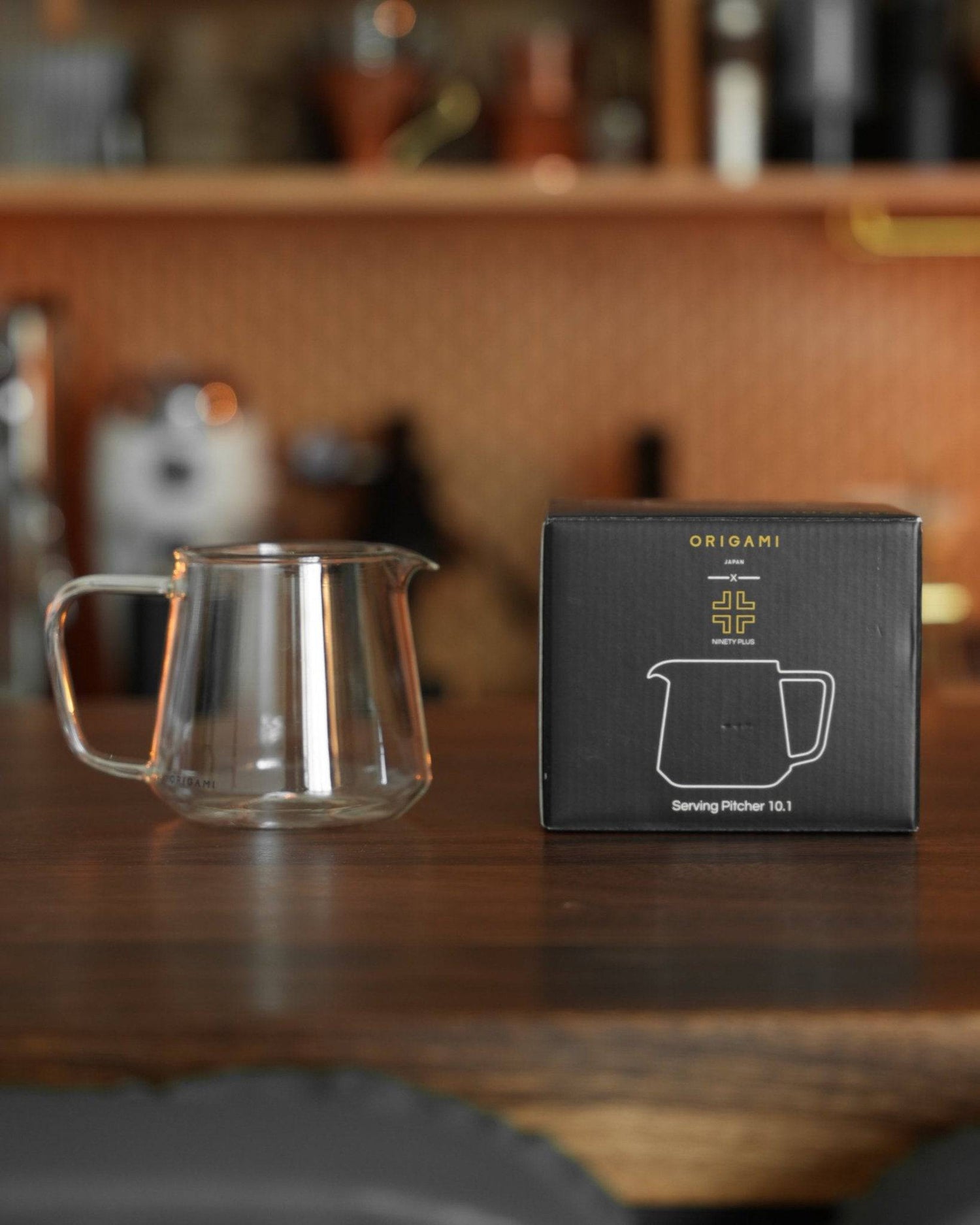 Ninety Plus Coffee Serving Pitcher 10.1 displayed on wooden counter next to its elegant black packaging featuring gold Origami logo, showcasing premium glassware for artisanal coffee brewing and serving