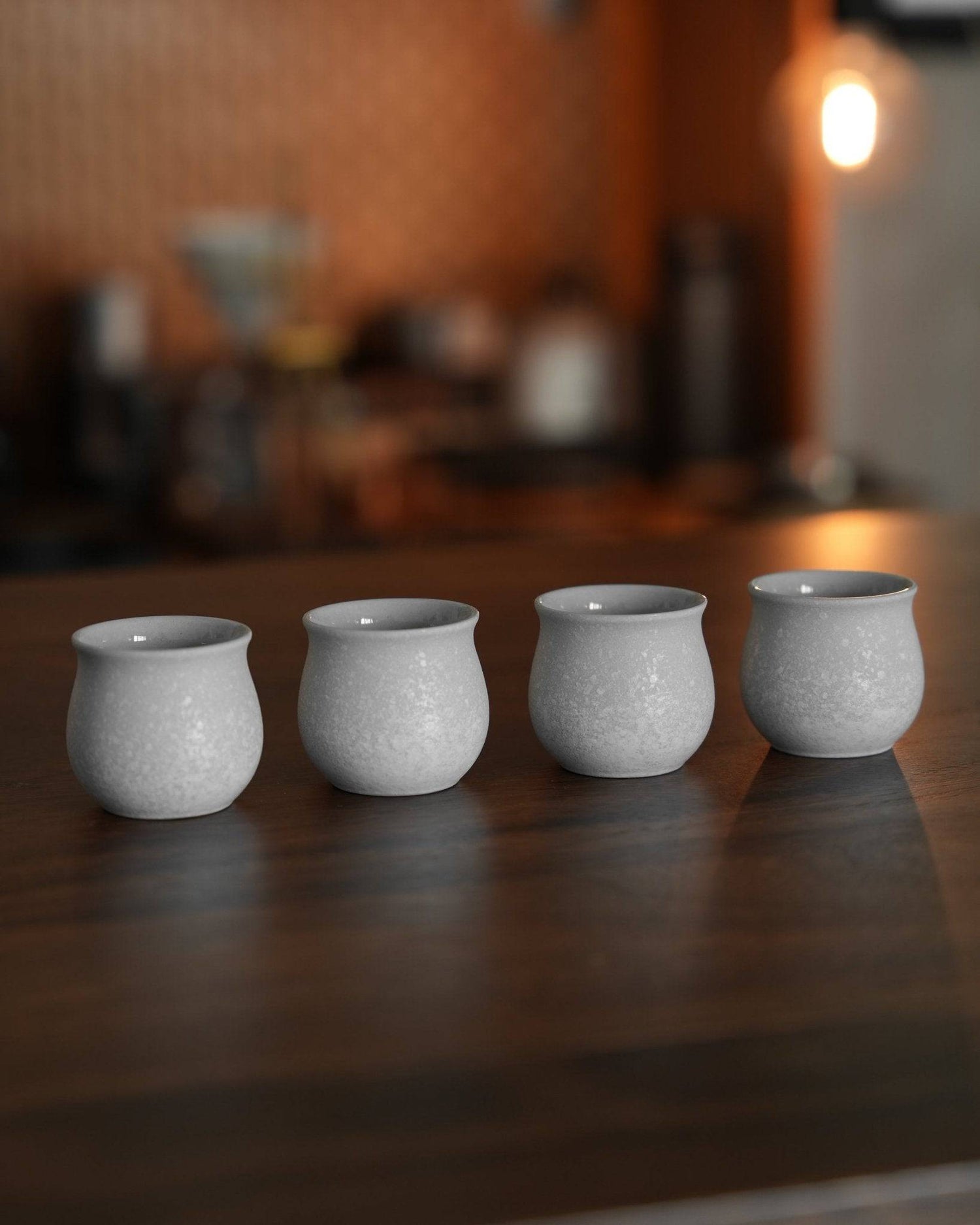 Four white ceramic Ninety Plus Coffee Sharing Cups on wooden table, designed for precise 2.5g coffee portions. Elegant set for sharing premium coffee experiences in a cafe setting with warm lighting and coffee equipment in background.