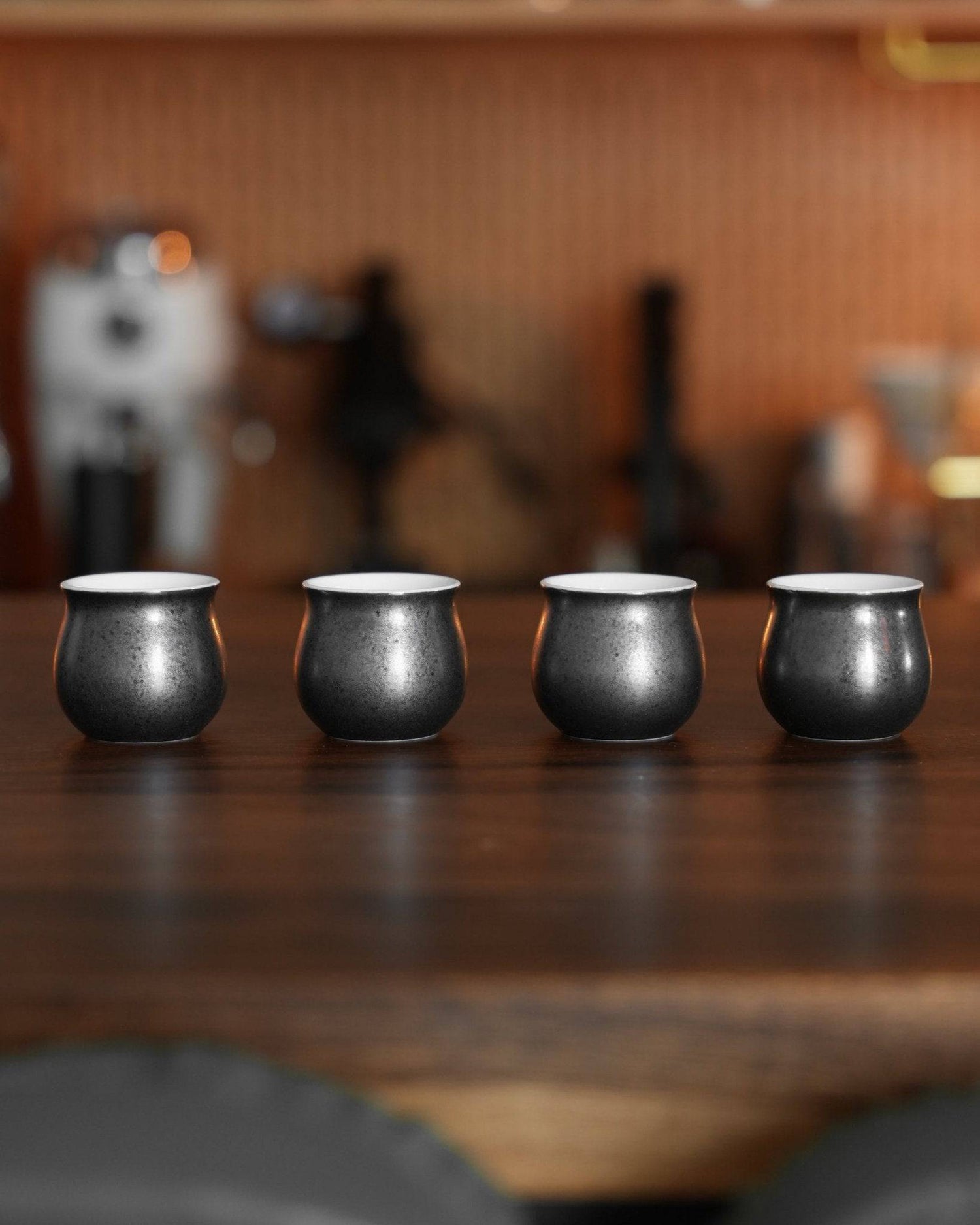 Four elegant black obsidian coffee sharing cups from Ninety Plus Coffee set on a wooden surface, with coffee equipment blurred in the background, showcasing the unique design for precise measurement and shared coffee experiences.