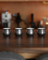 Four elegant black obsidian coffee sharing cups from Ninety Plus Coffee set on a wooden surface, with coffee equipment blurred in the background, showcasing the unique design for precise measurement and shared coffee experiences.