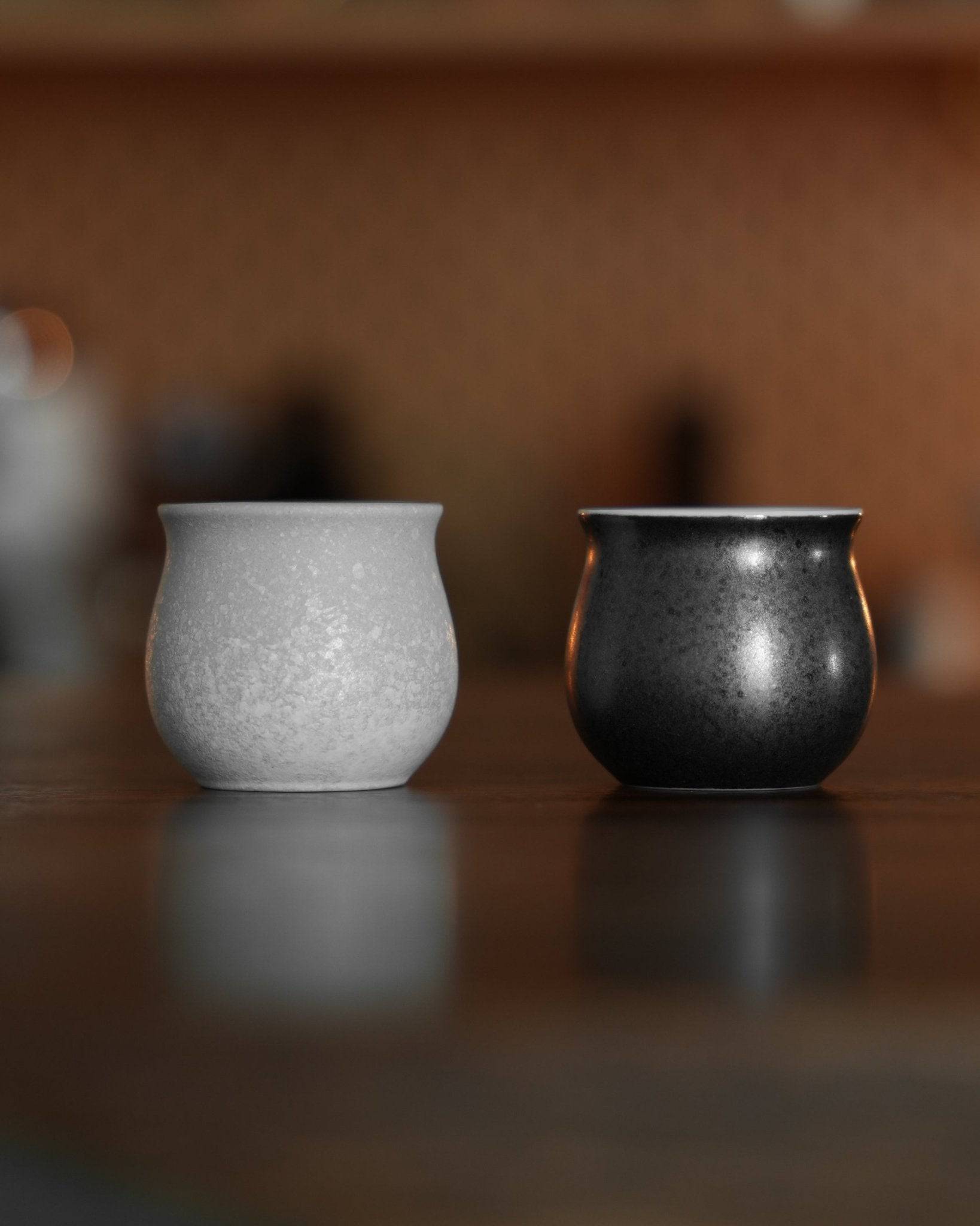 Two small, rounded ceramic cups side by side on a reflective surface. One cup is white with a speckled texture, the other is black with a glossy finish. Both have a slightly bulbous shape, ideal for serving specialty coffee.