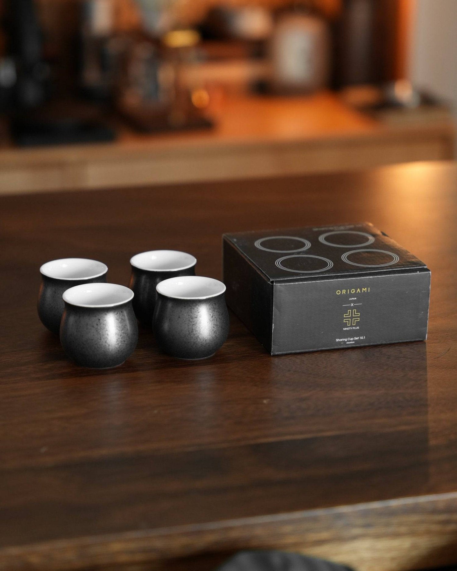 Ninety Plus Coffee Sharing Cup Set featuring four black ceramic cups with white interiors and a sleek black packaging box, displayed on a wooden surface in a cafe setting