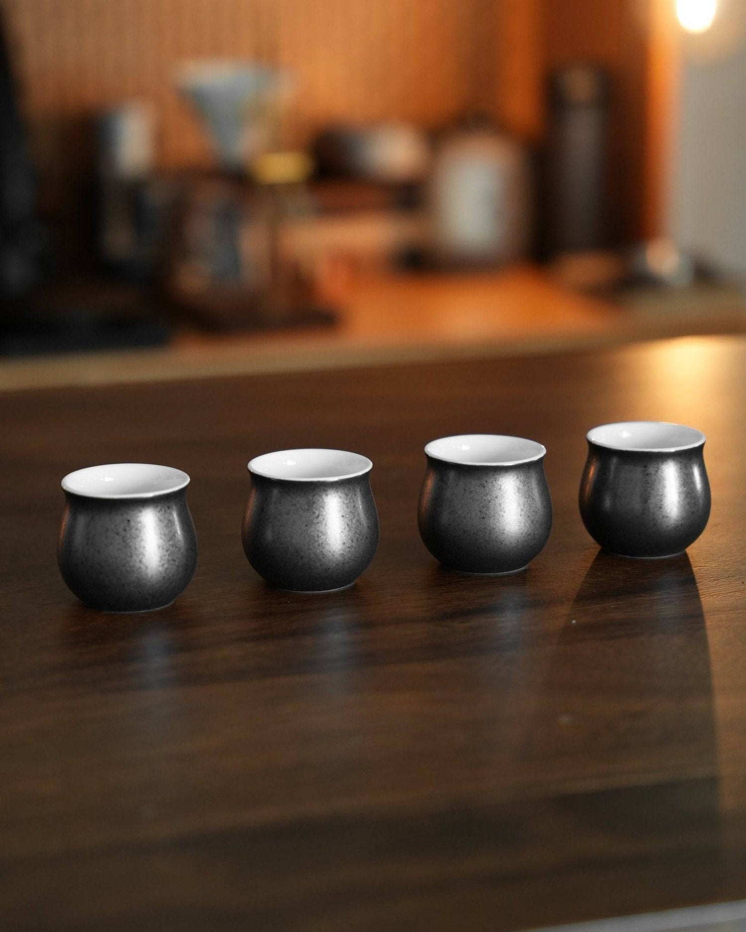 Four sleek black obsidian coffee sharing cups from Ninety Plus Coffee set on a wooden counter, with blurred coffee equipment in the background, showcasing elegant design for premium coffee tasting experience.