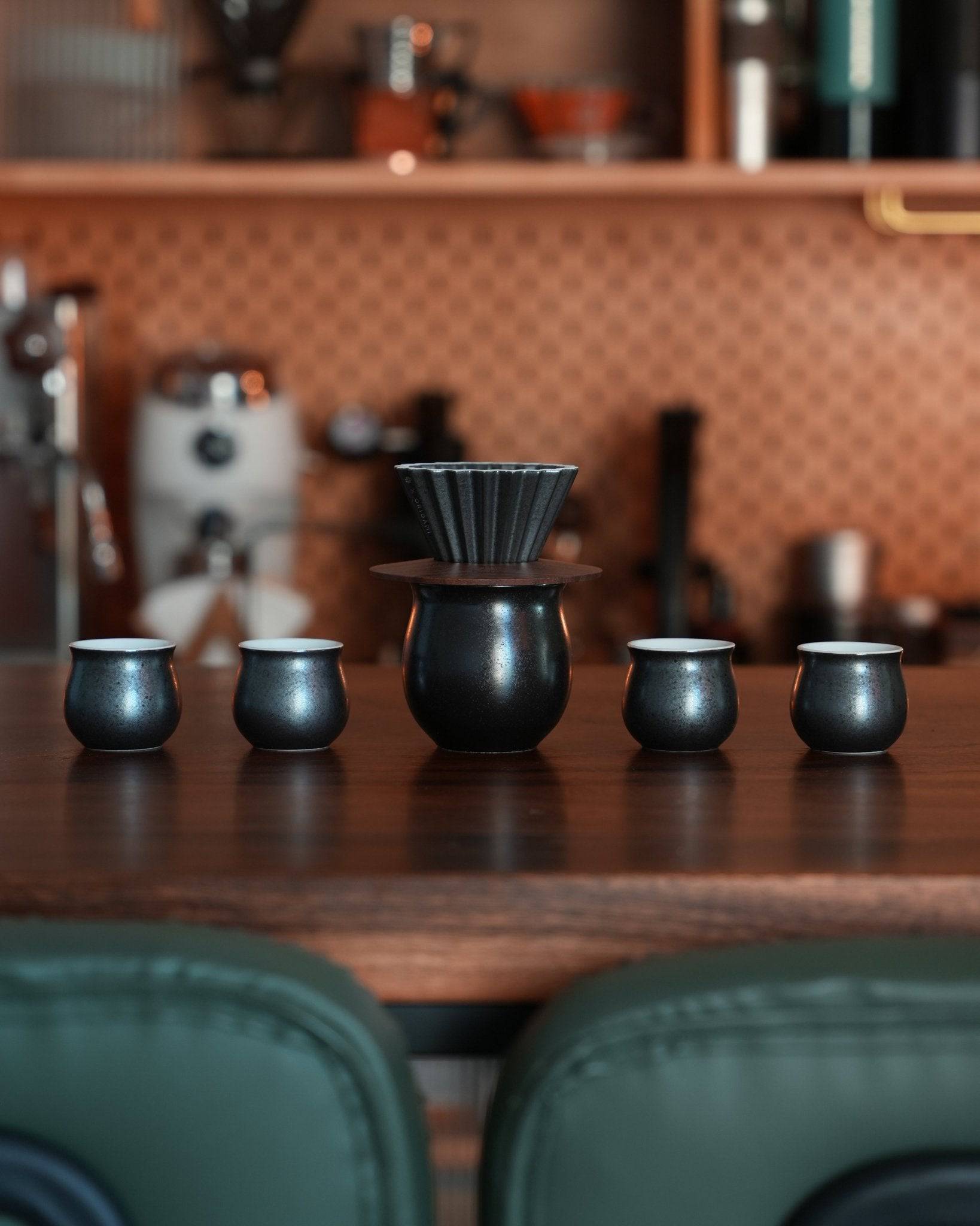 Ninety Plus pour-over coffee set in obsidian black, featuring a ceramic dripper and four matching cups, displayed on a wooden counter in a stylish coffee shop setting with copper-toned backsplash and green chairs visible