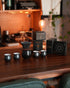 Ninety Plus pour-over coffee set in elegant black, featuring dripper, cups, and product boxes arranged on wooden table in warm, cozy kitchen setting with copper backsplash and ambient lighting