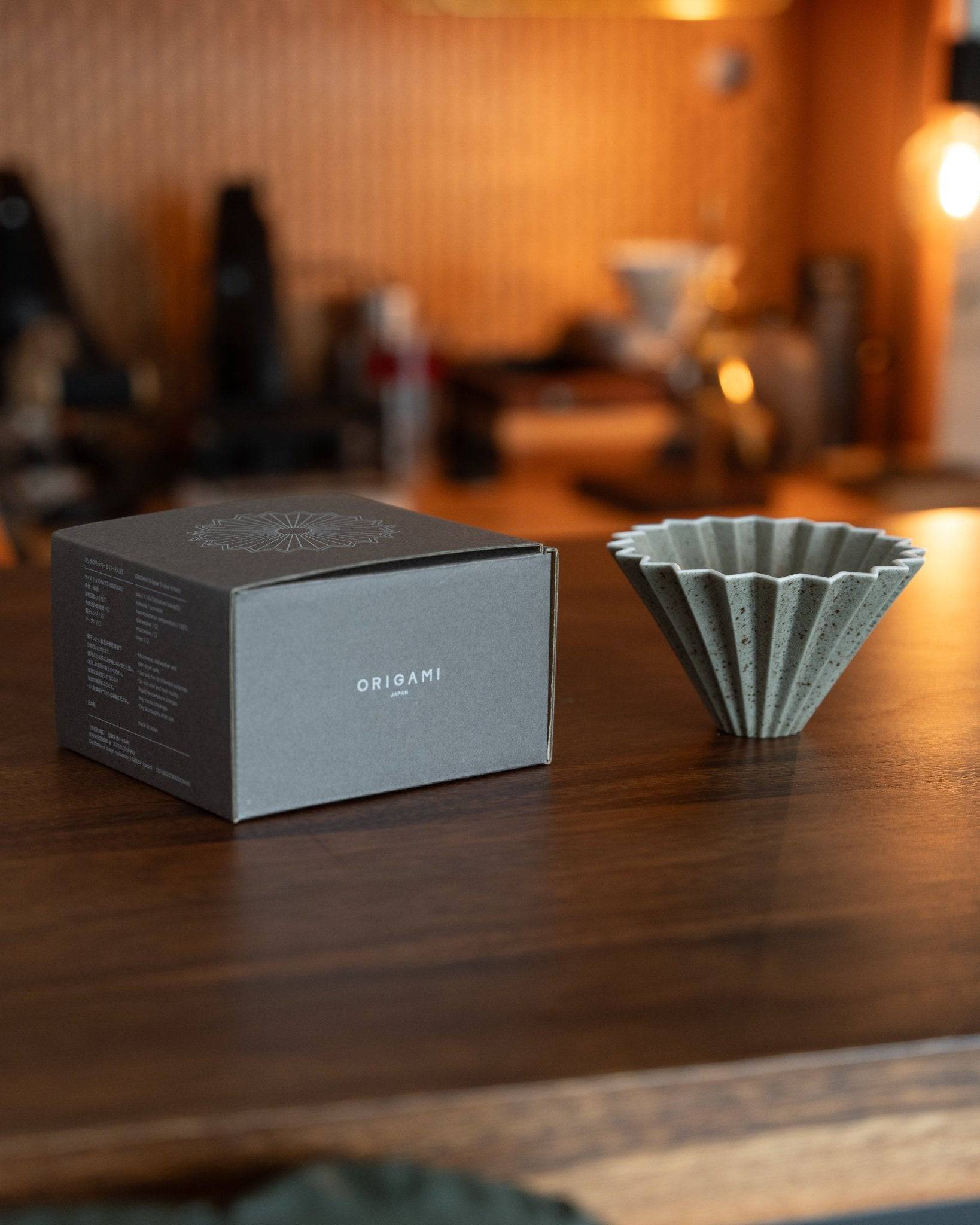 Origami Dripper Mikage limited edition coffee filter with unique sand glaze texture, displayed next to its packaging box on a wooden surface in a warm, coffee shop-like setting