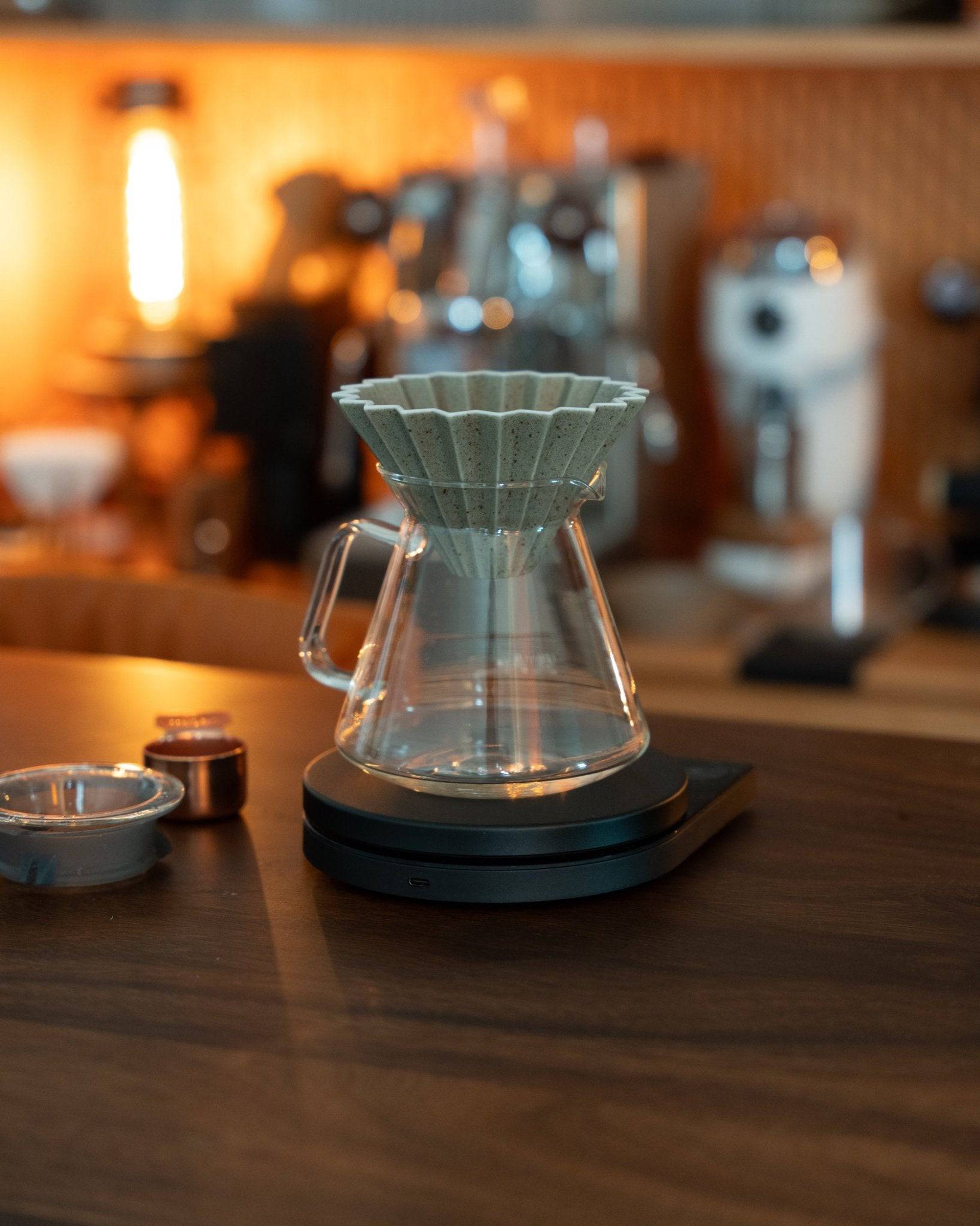Origami Dripper Mikage set on wooden counter with coffee equipment in background, showcasing unique pour-over design and Japanese craftsmanship in warm, inviting cafe ambiance