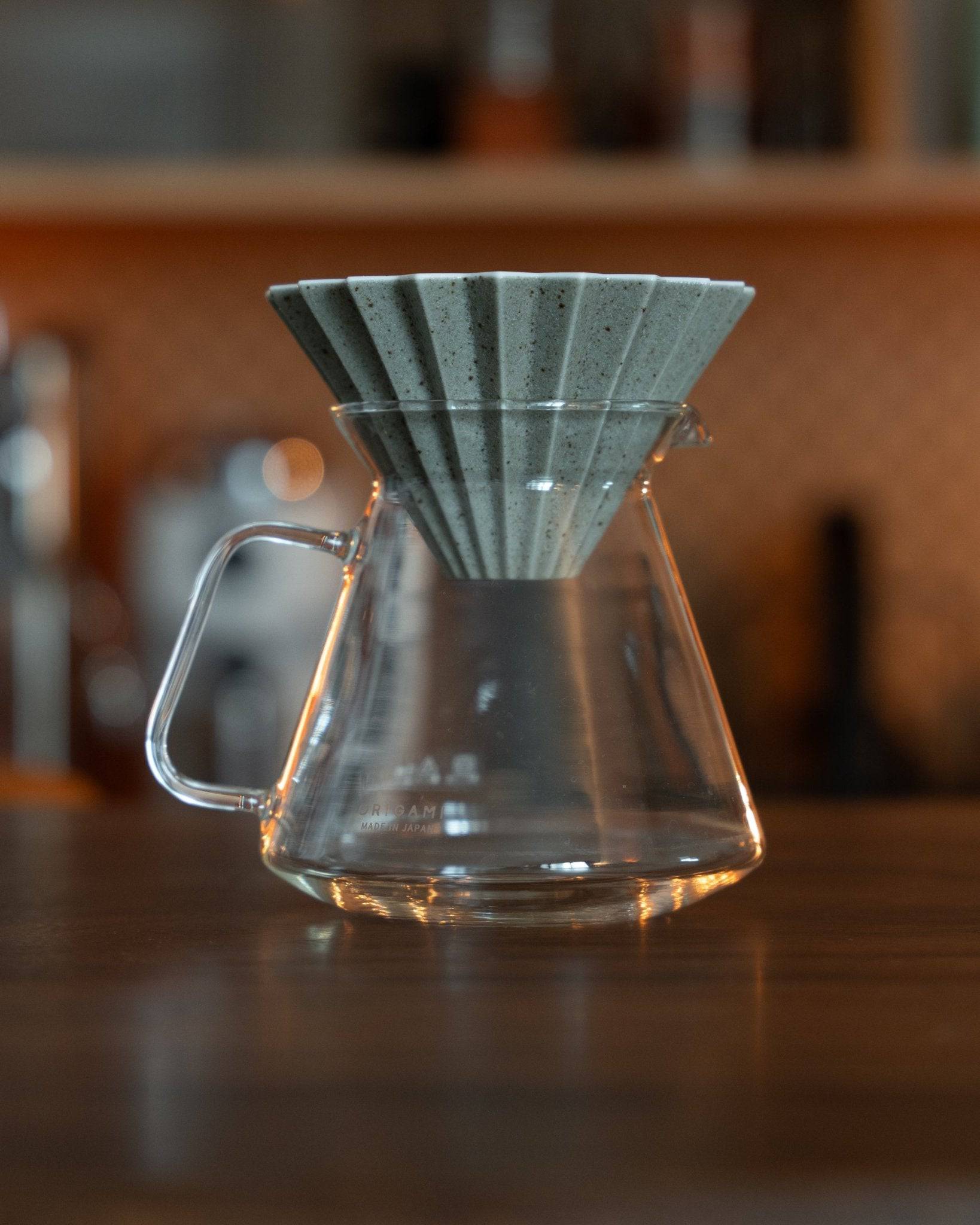 Origami Dripper Mikage sand glaze limited edition pour-over coffee filter on glass carafe, showcasing unique texture and color, embodying Japanese wabi-sabi aesthetics, on wooden counter with blurred background
