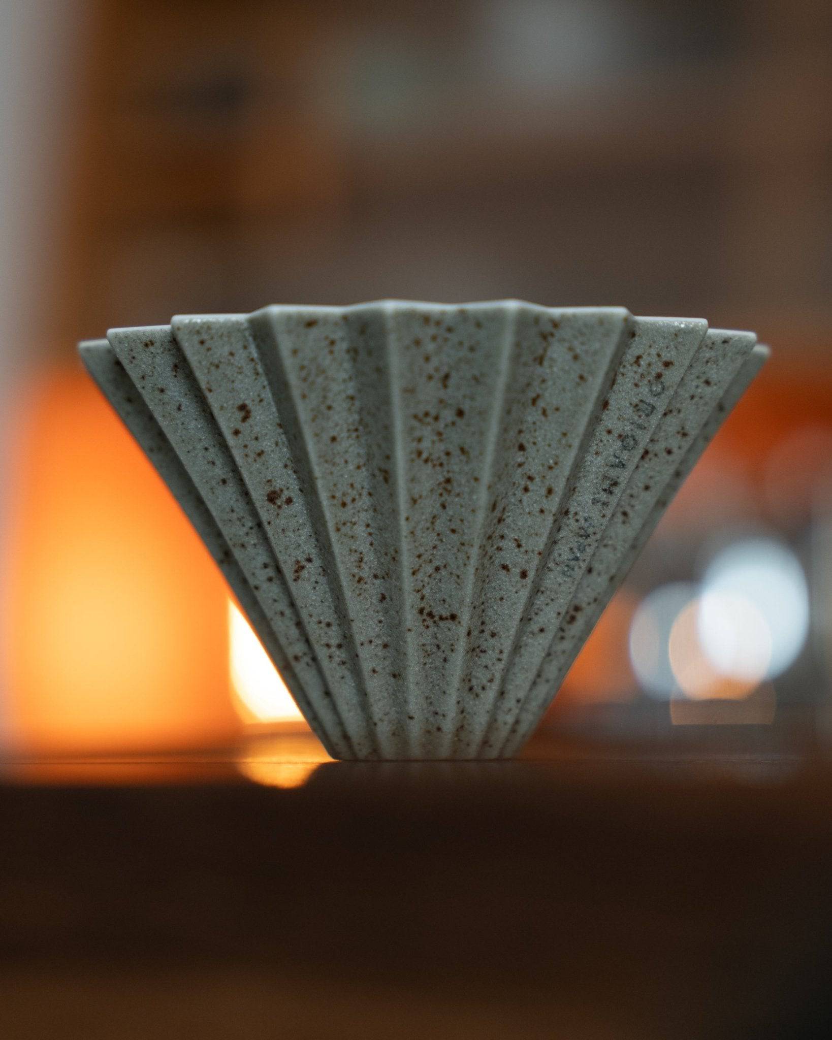 Origami Dripper Mikage limited edition coffee filter, featuring unique sand-glaze texture and speckled pattern. Conical shape with ribbed exterior. Warm orange glow in background, showcasing the ceramic&