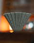Origami Dripper Mikage limited edition coffee filter, featuring unique sand-glaze texture and speckled pattern. Conical shape with ribbed exterior. Warm orange glow in background, showcasing the ceramic&