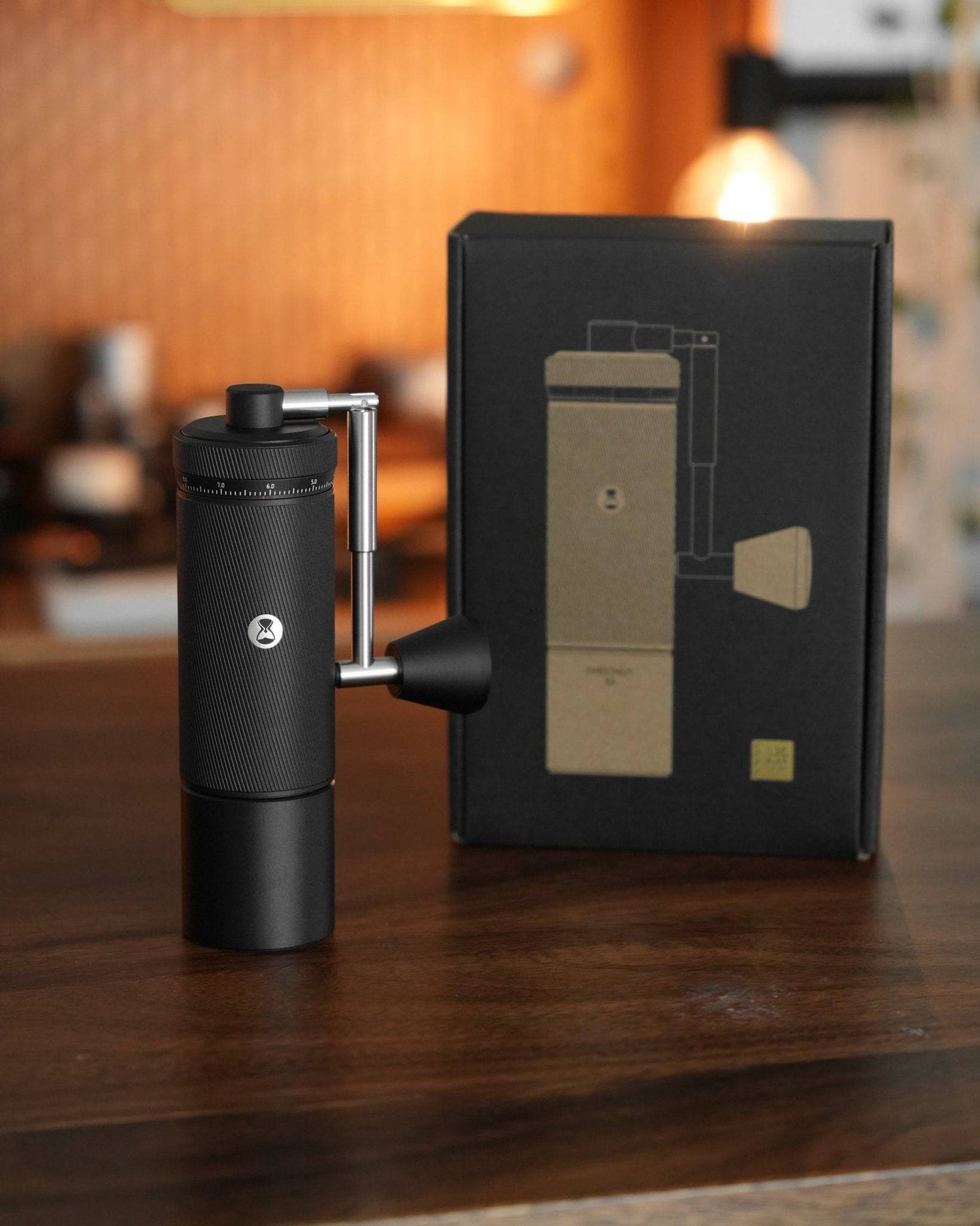 Timemore Chestnut S3 manual coffee grinder with external adjustment dial, displayed next to its sleek black packaging on a wooden table in a warm, cozy setting