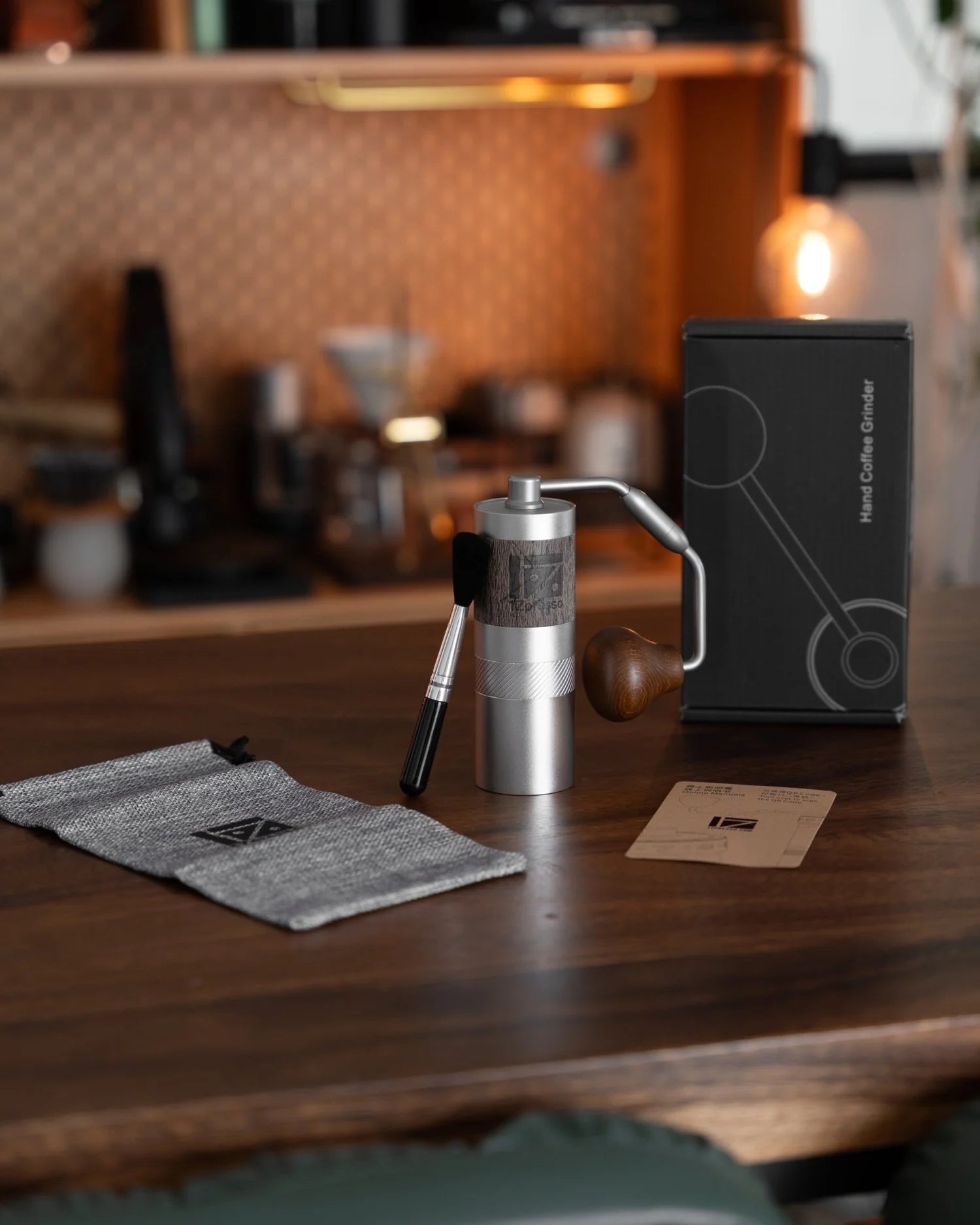 1ZPresso Q portable coffee grinder on wooden table with accessories, packaging, and storage bag in warm, cozy kitchen setting. Sleek design showcases premium materials and compact size for travel and outdoor use.