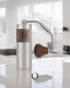 1ZPresso Q portable coffee grinder with silver metal body and wooden accent, featuring ergonomic handle and adjustable grind settings, displayed on a white kitchen counter in a bright, modern interior.