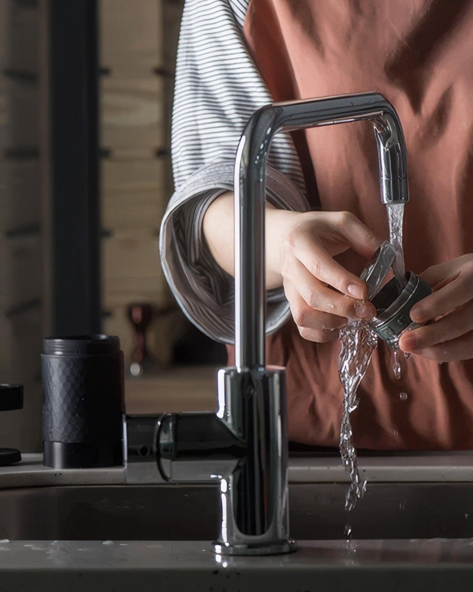 Hands rinsing a coffee brewing device under a modern kitchen faucet, with a sleek black coffee grinder visible in the background, showcasing the coffee preparation process