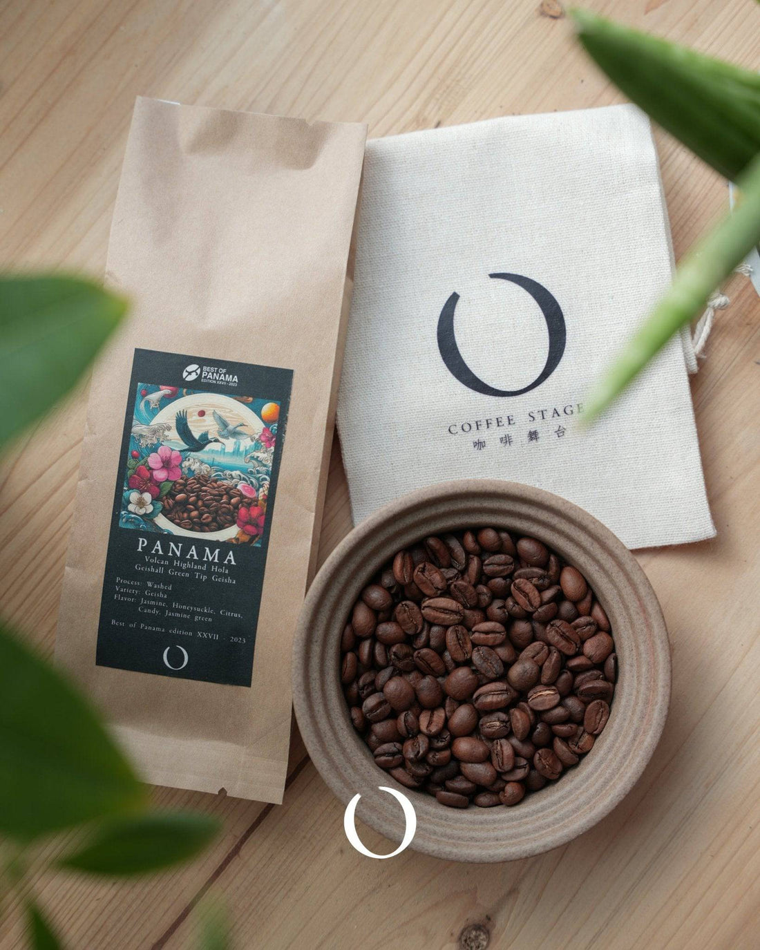 Premium Panama Geisha coffee beans in a ceramic bowl, alongside branded packaging and a Coffee Stage cloth, on a wooden surface with green leaves in view