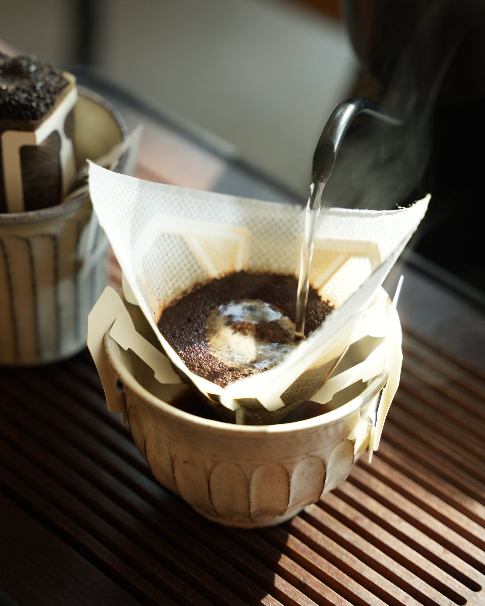 Hot water being poured over ground coffee in a drip filter bag, placed in a ceramic cup on a wooden surface, demonstrating the pour-over brewing method for Yunnan CATIMOR 14A light roast coffee.