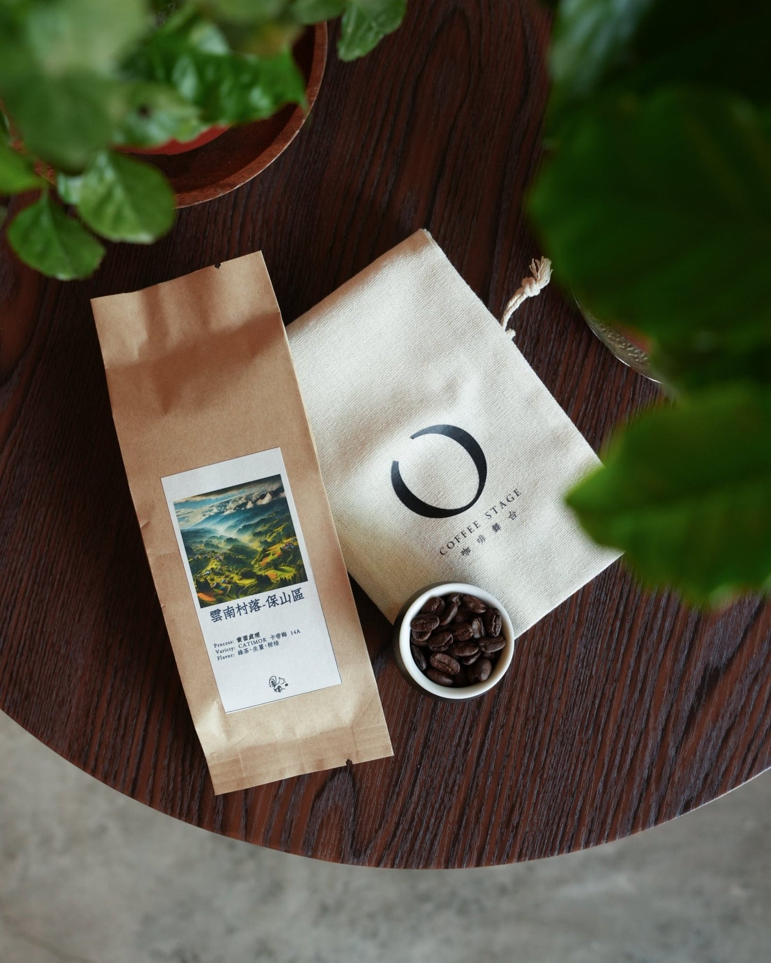 Coffee packaging from Yunnan village displayed on wooden table with branded cloth bag and small bowl of coffee beans, surrounded by green plant leaves