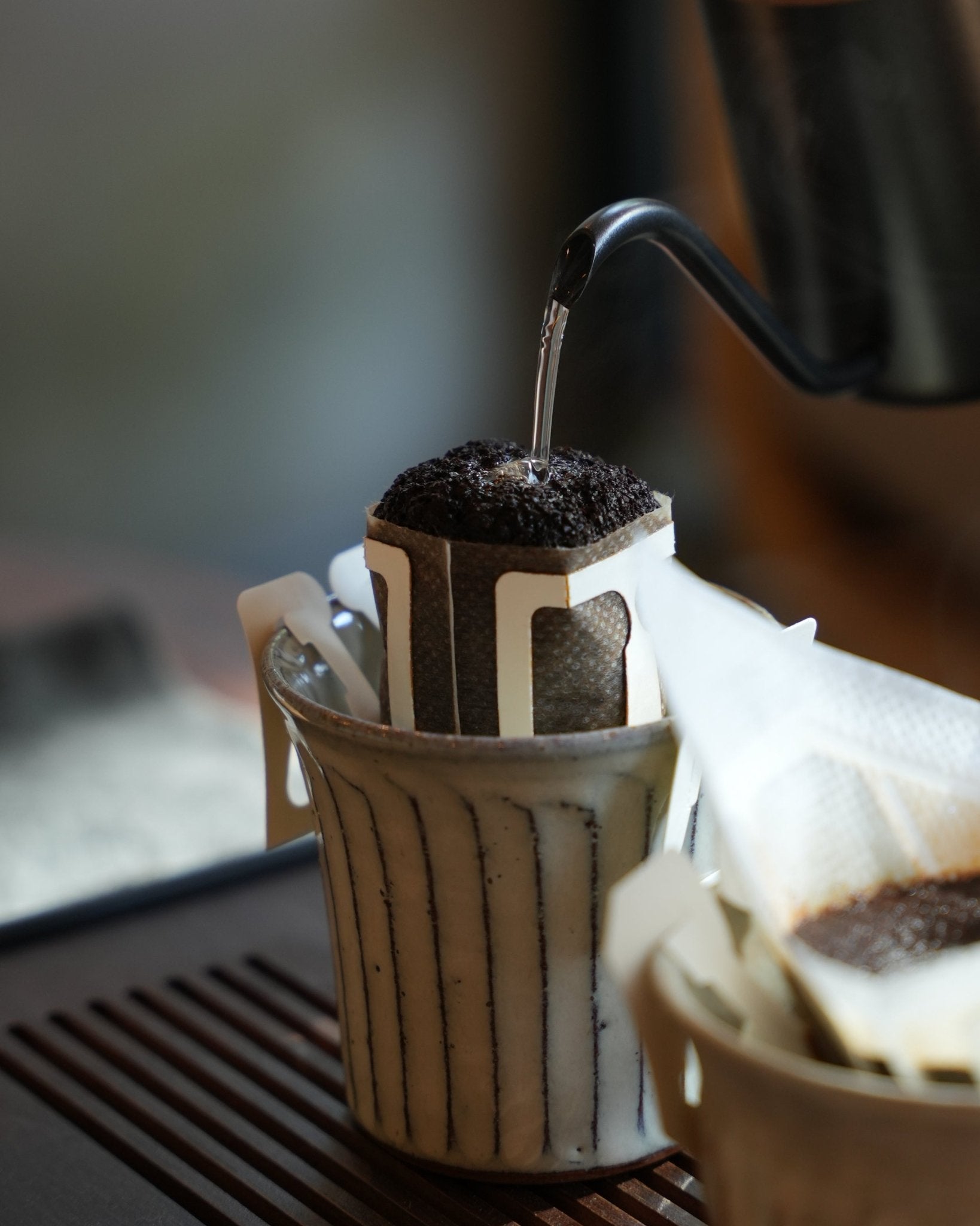 Close-up of a drip coffee bag in a ceramic cup, with hot water being poured from a kettle spout. The scene showcases the process of brewing Yunnan Catimor 14A coffee using the pour-over method.