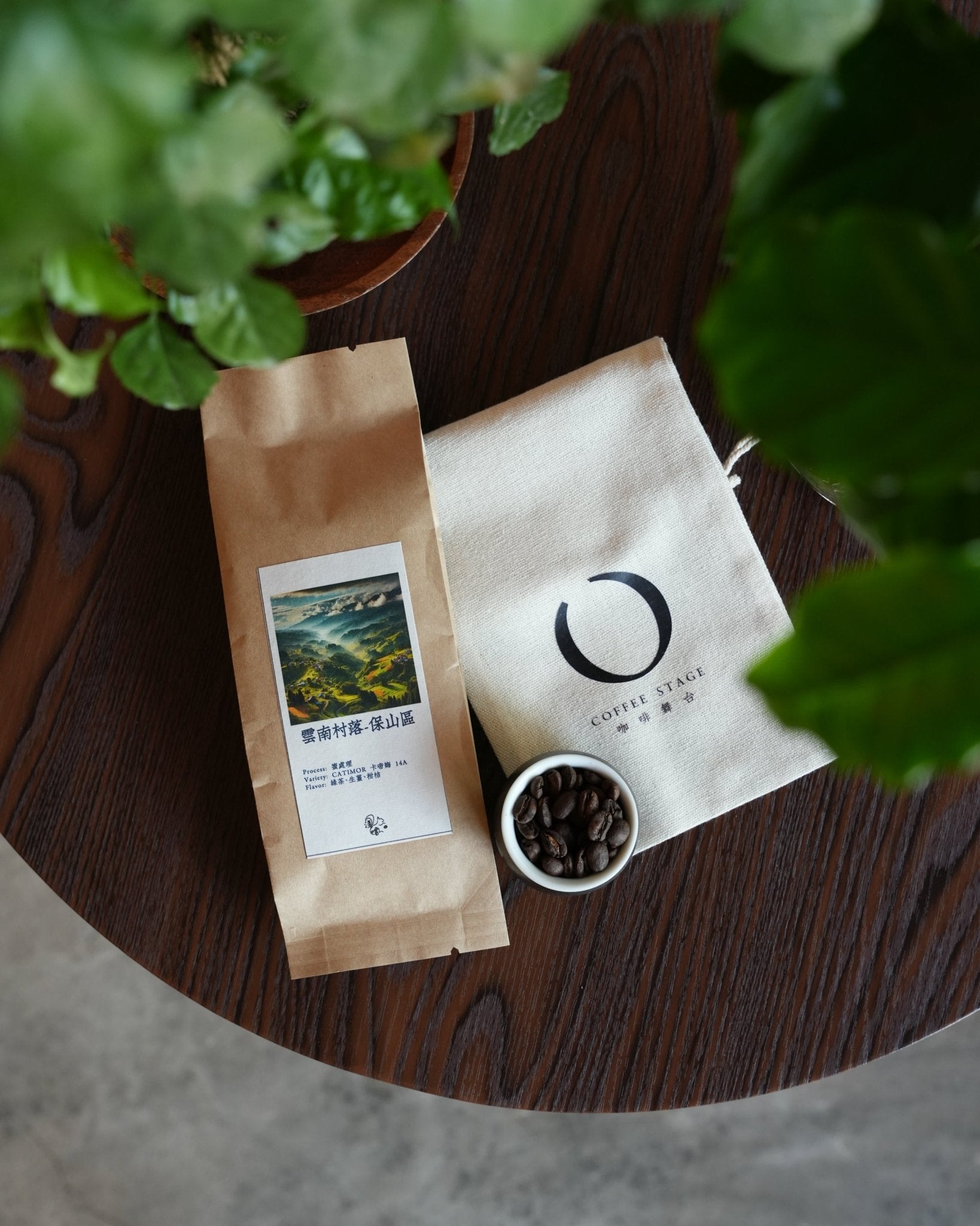 Artfully arranged coffee product display featuring a brown paper bag of Yunnan coffee beans, a white canvas pouch with logo, and a small bowl of roasted coffee beans on a wooden surface, framed by green leaves.