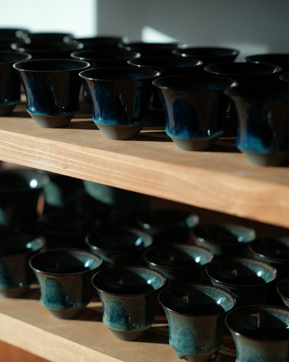 Finished cups arranged on a workshop rack, their dark-blue glaze reflecting window light