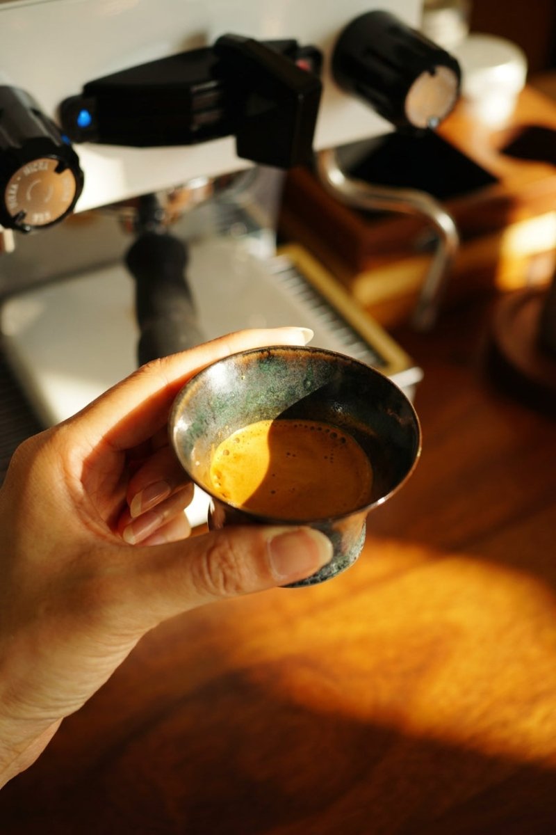 手持 Coffee Stage Sharing Cup S Size，杯內盛有少量 Espresso，背景為咖啡機