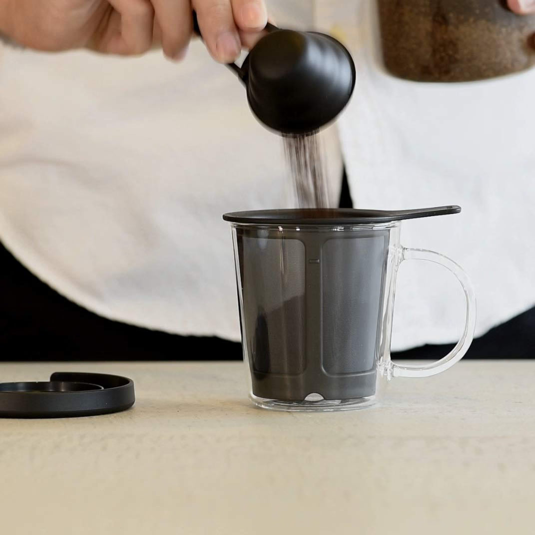 HARIO One Cup 單杯網濾咖啡器 170mL：咖啡粉正倒入 One Cup 內置聚酯纖維濾網，杯蓋放在咖啡器旁。