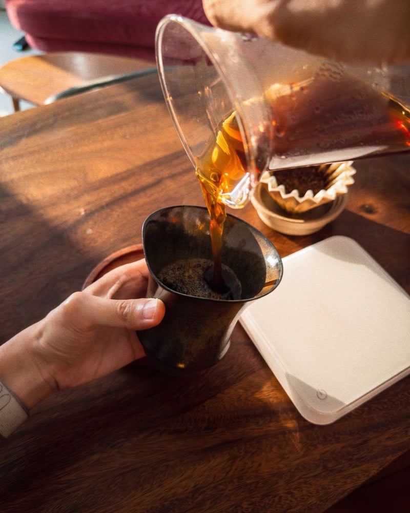 手沖咖啡從玻璃分享壺注進燼金黑釉的 Alpha 杯，旁邊有濾杯與濾紙