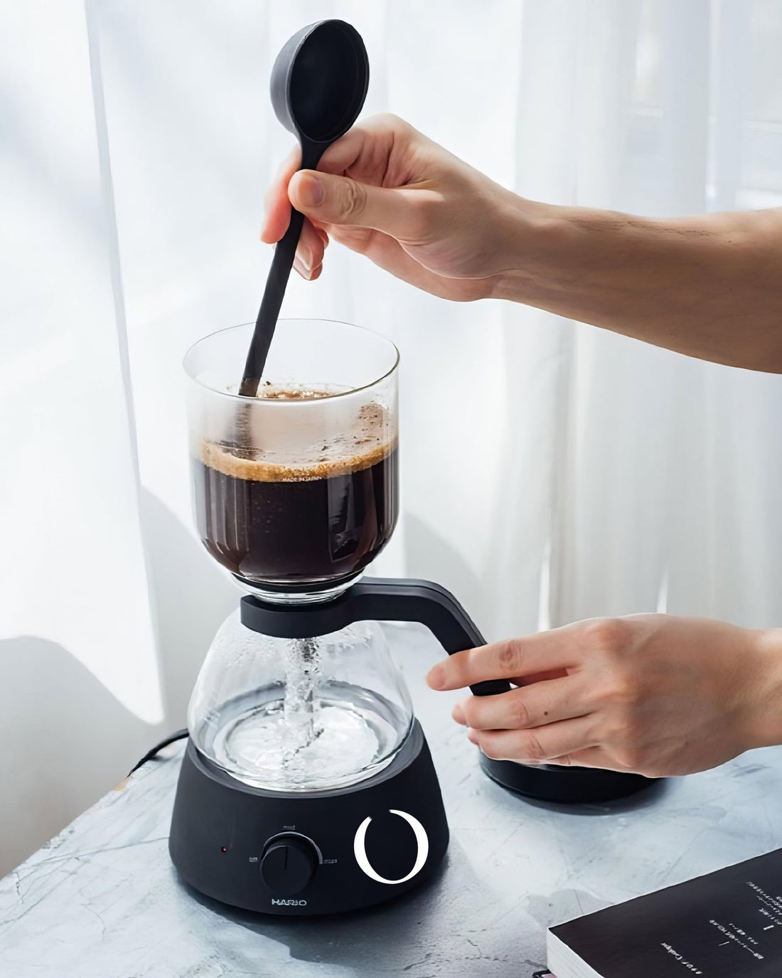 HARIO electric siphon coffee maker in use, showing hands stirring coffee in upper glass chamber, with lower chamber on heating base, demonstrating the brewing process of siphon coffee