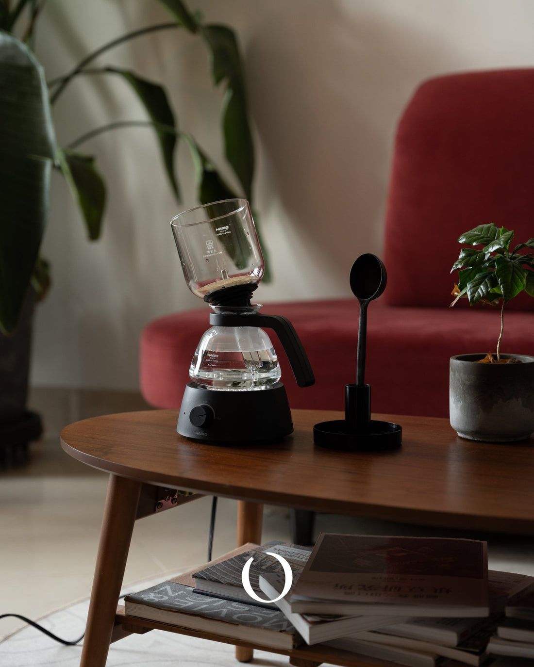 HARIO electric siphon coffee maker on wooden table with red chair and plants, creating a cozy home coffee brewing setup