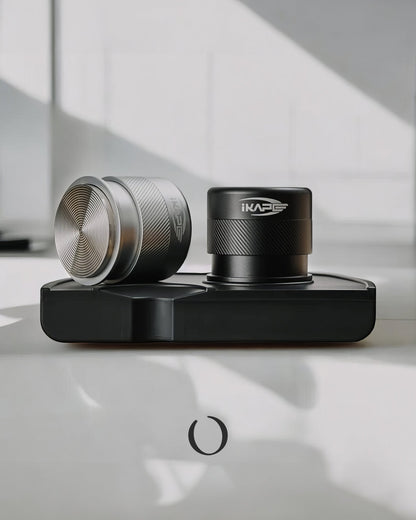 IKAPE Espresso coffee tampers in silver and black, featuring calibrated spring design for consistent pressure, displayed on a sleek black stand against a white surface with shadow patterns