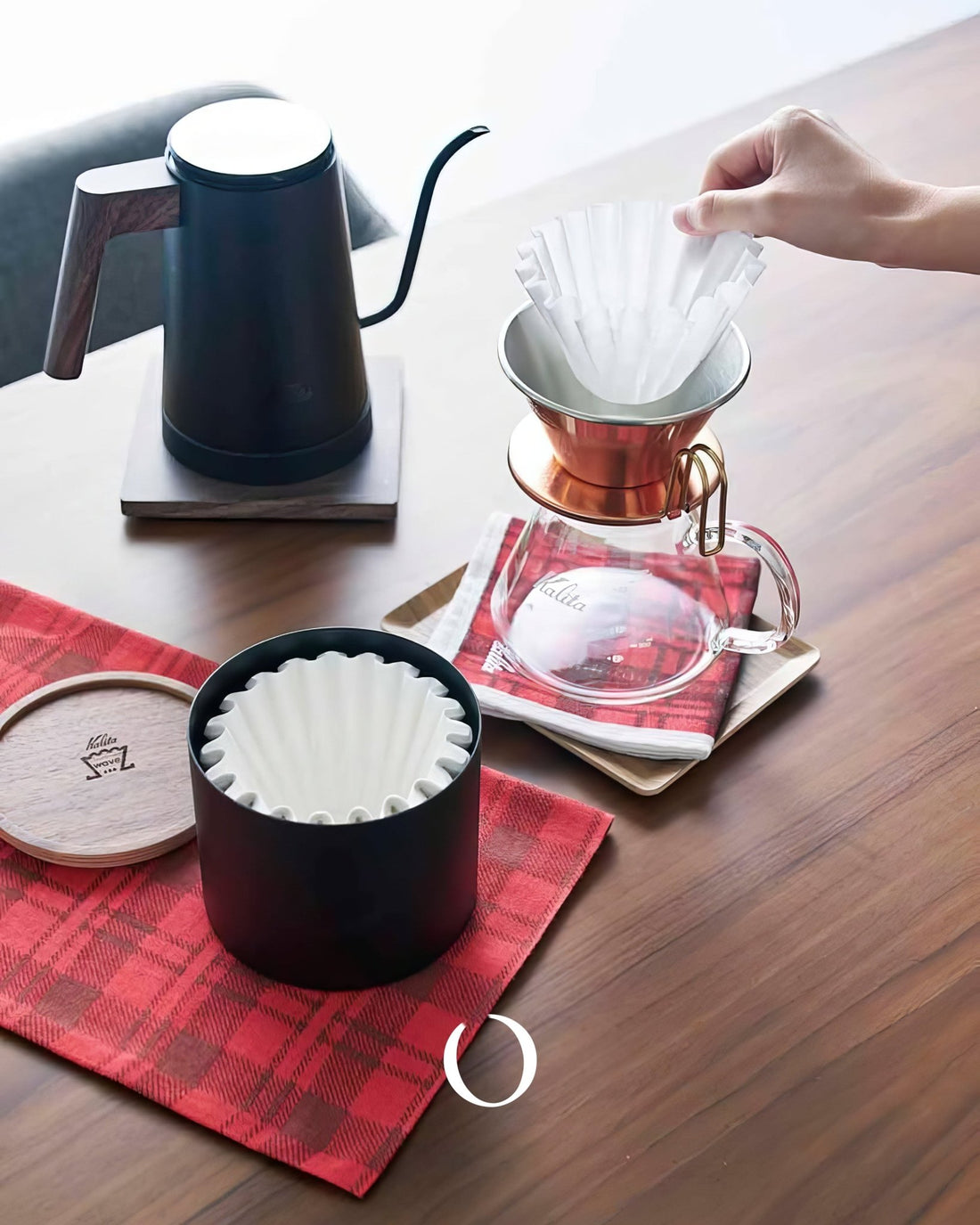 Kalita coffee brewing setup featuring gooseneck kettle, Kalita Wave dripper, glass server, and Roshi filter paper container on wooden table with red plaid cloth, showcasing pour-over coffee preparation process