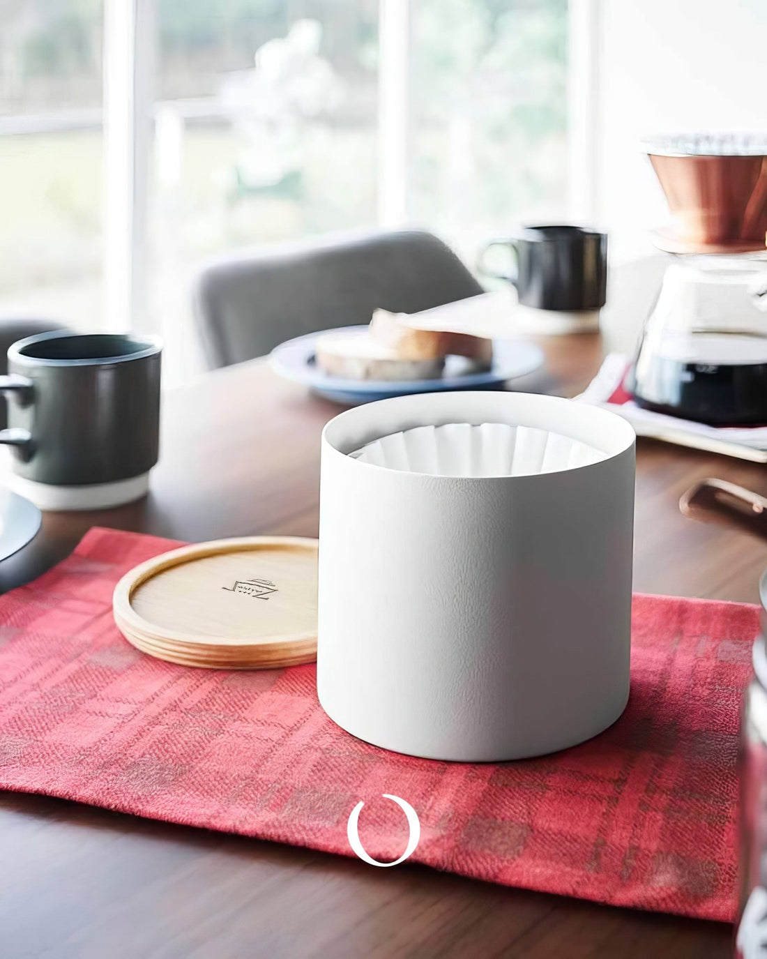 Kalita 155/185 Roshi filter paper container on red cloth, surrounded by coffee brewing equipment on wooden table near window, showcasing product in use with coffee maker, mugs, and bread plate in background