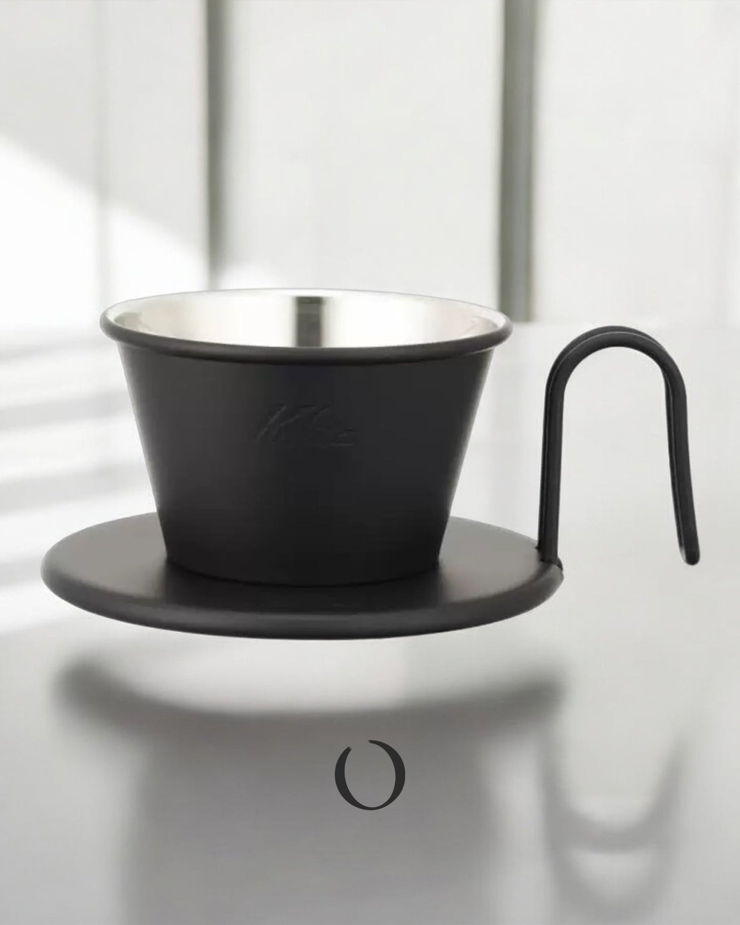 Limited edition Kalita Wave Dripper in matte black finish with stainless steel interior, sitting on a black coaster with curved handle, showcasing its elegant design for pour-over coffee brewing