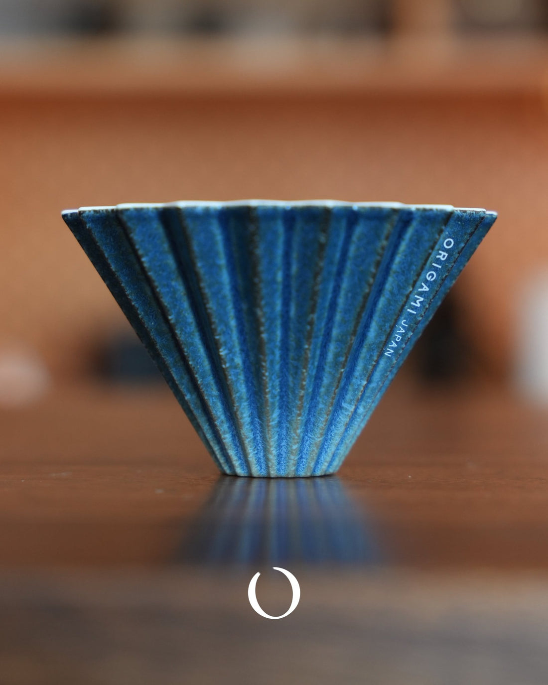 Limited edition ORIGAMI Dripper S [sukumo] coffee filter, blue ceramic with unique indigo-dyed pattern, ribbed design for optimal brewing, displayed on wooden surface, showcasing Japanese craftsmanship and coffee culture fusion