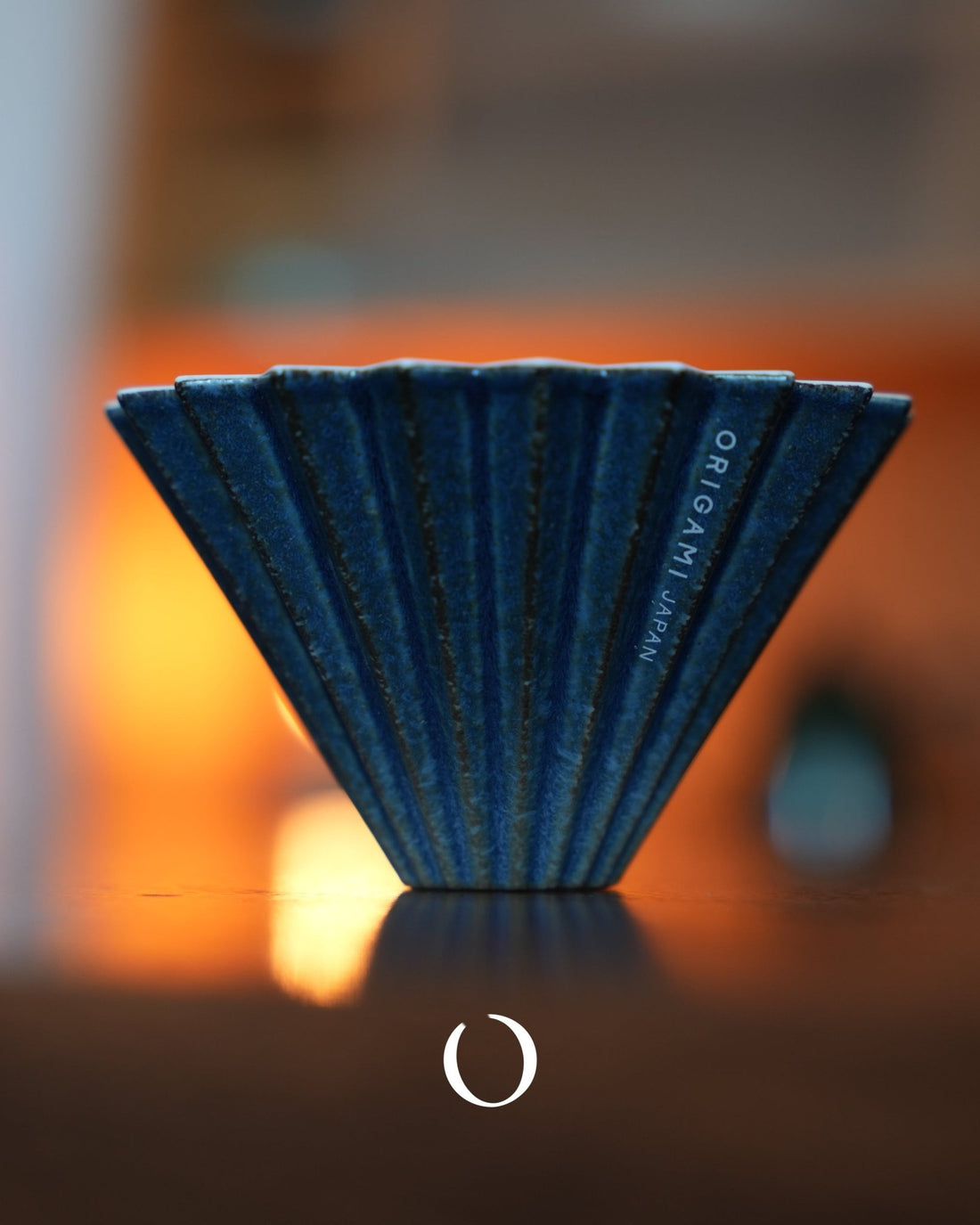 Limited edition ORIGAMI Dripper S [sukumo] coffee filter, featuring unique blue indigo-dyed ceramic with ribbed design, showcased against warm background lighting, highlighting its artisanal craftsmanship and exclusive nature