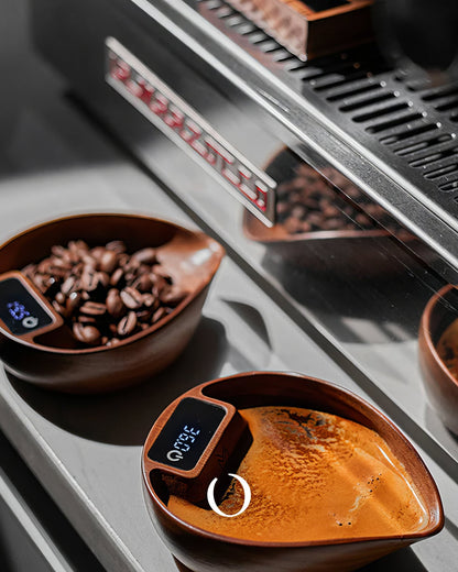 MUVNA walnut wood coffee scale with digital display, showing coffee beans and brewed espresso in wooden bowls on a stainless steel surface next to an espresso machine